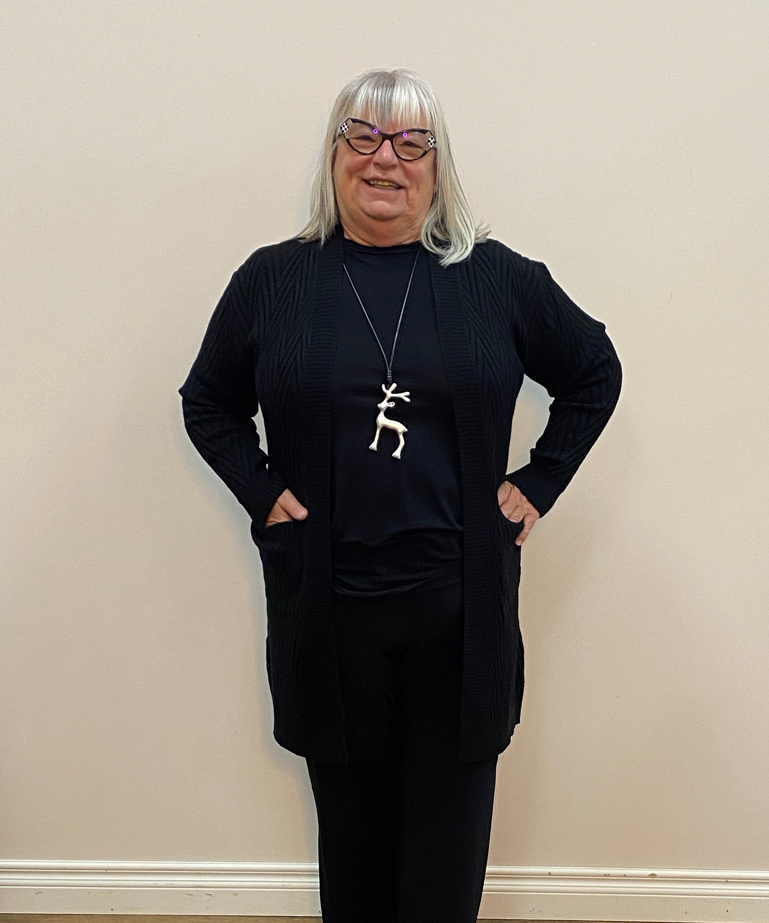 Person wearing a black outfit with a white necklace in a room with wooden flooring and a white wall.