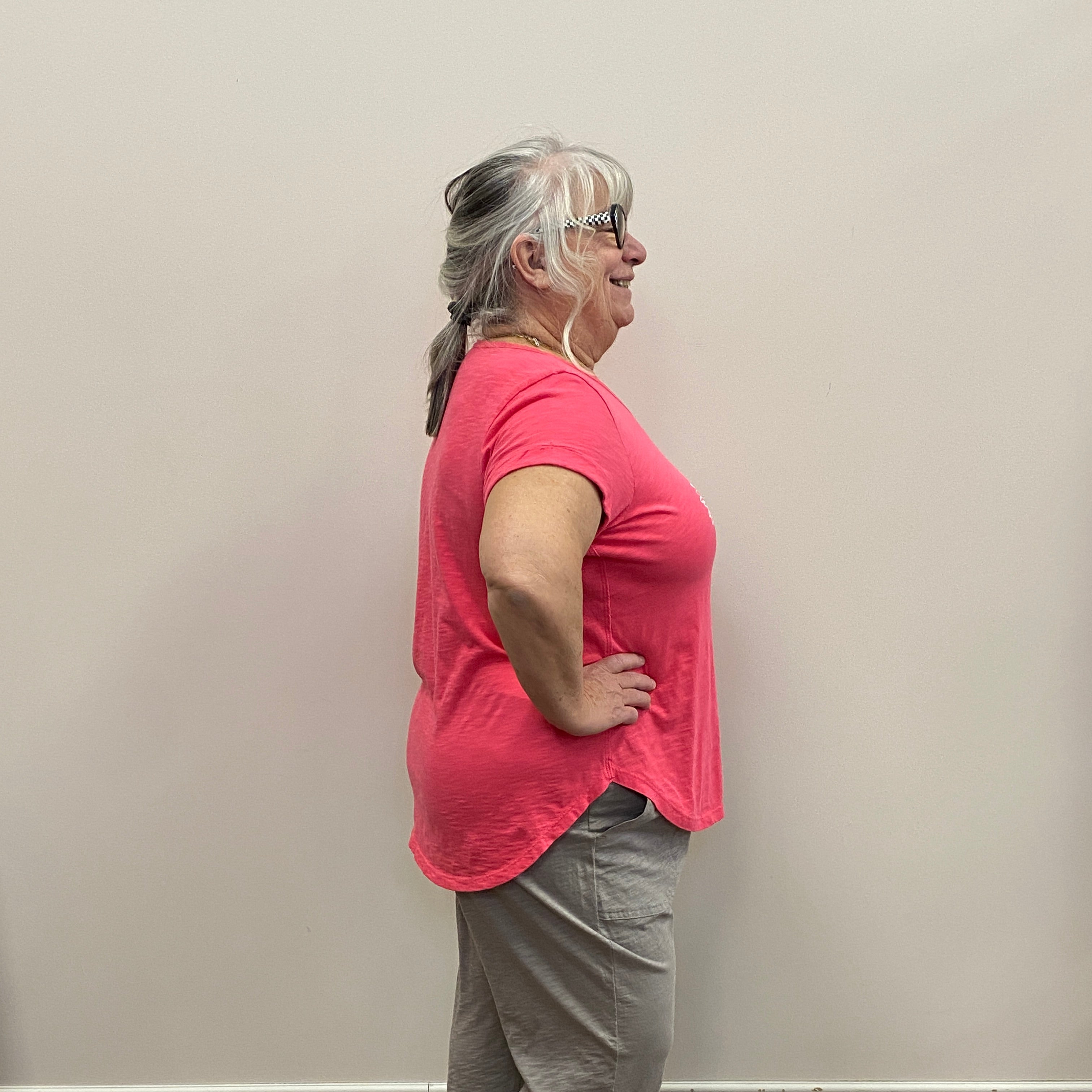 Person in a pink shirt and gray pants standing against a plain wall.