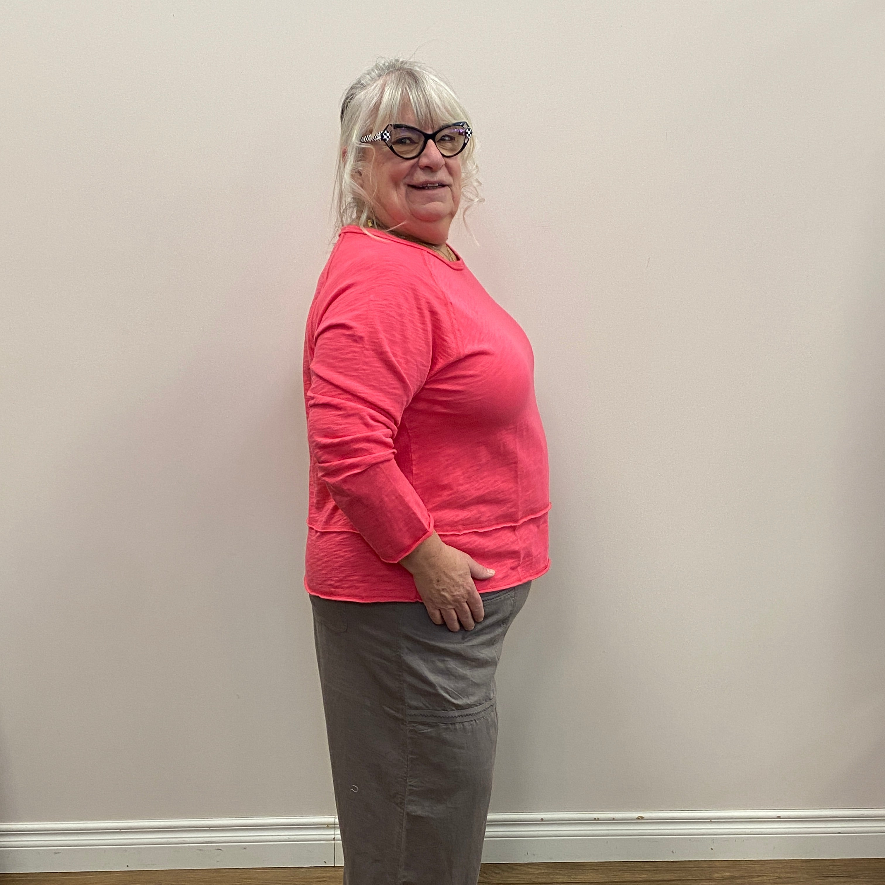 Person wearing a pink sweatshirt and gray pants standing against a white wall with wooden flooring.