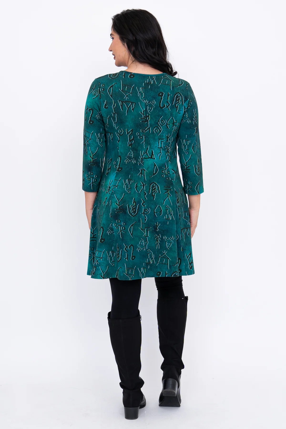 Woman wearing a teal patterned dress with black leggings and boots on a white background