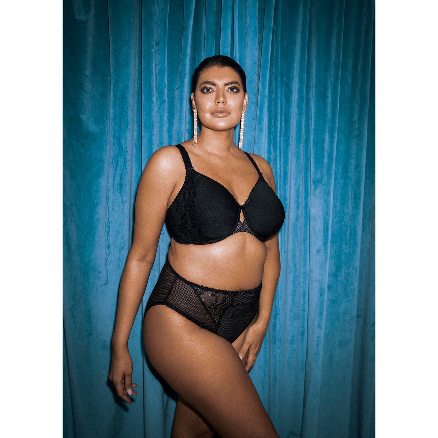 A woman wearing a black bra and panty set with lace details against a teal curtain.