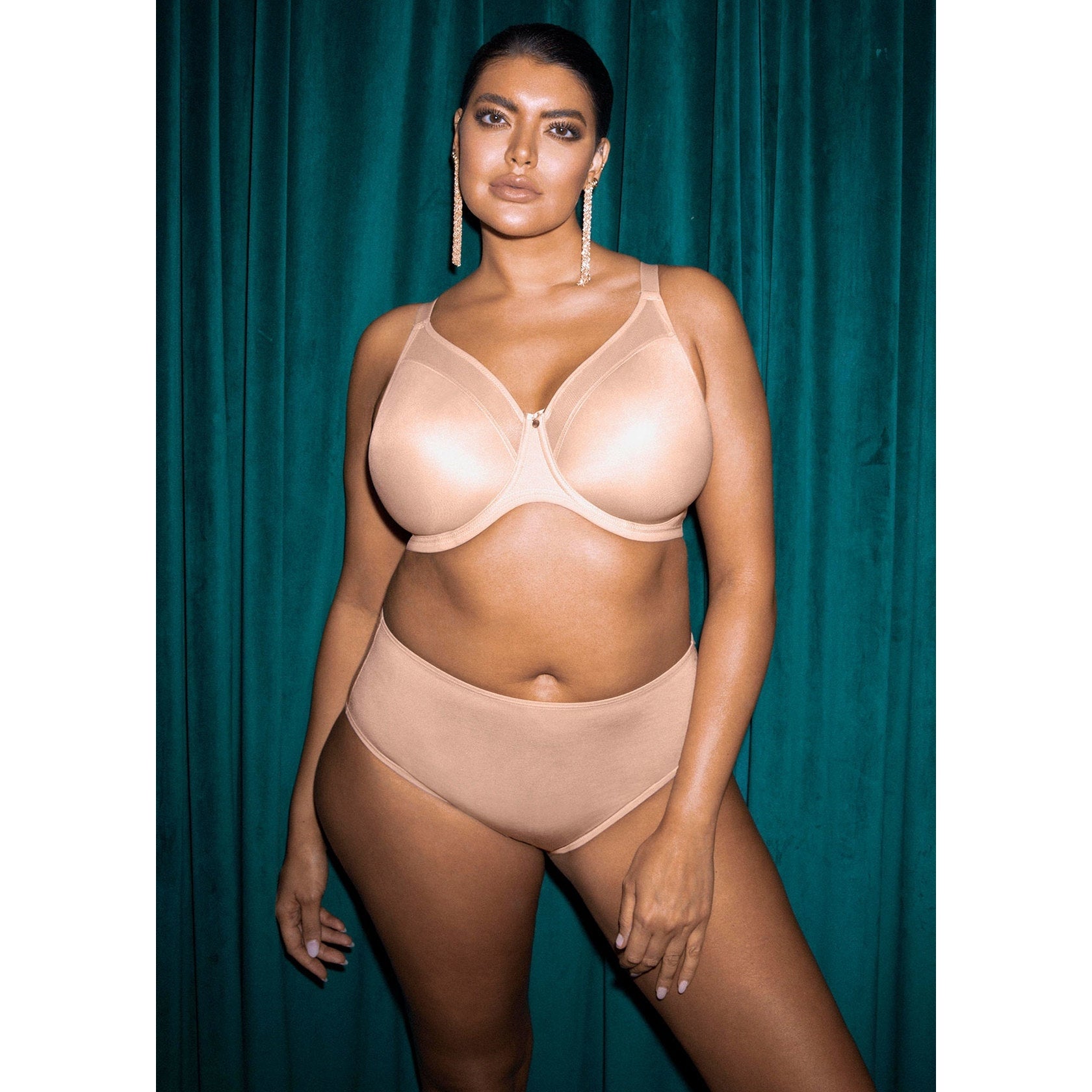A woman wearing a smooth beige bra and panty set against a teal curtain.