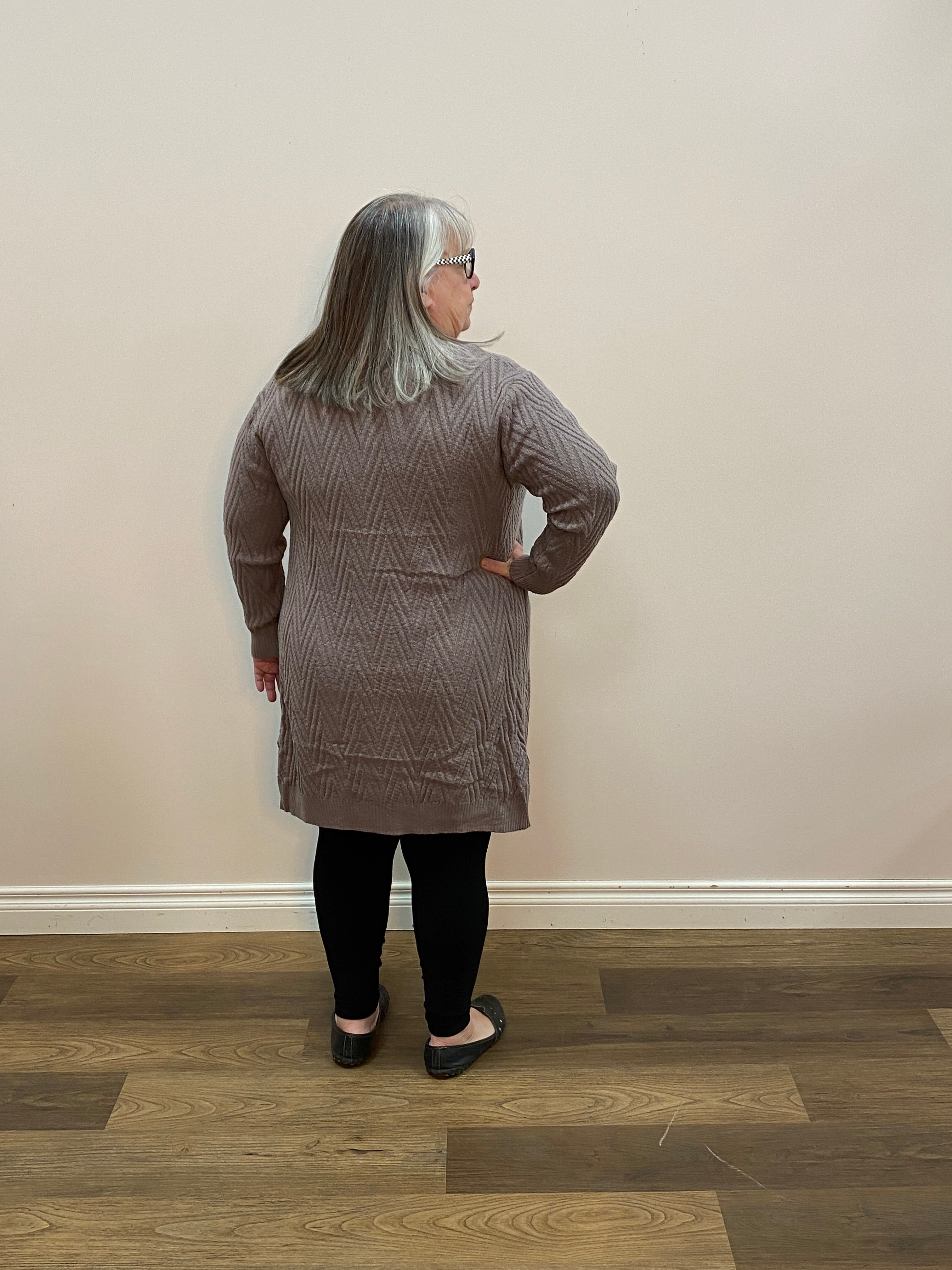 Person wearing a textured gray sweater and black pants standing on a wooden floor.