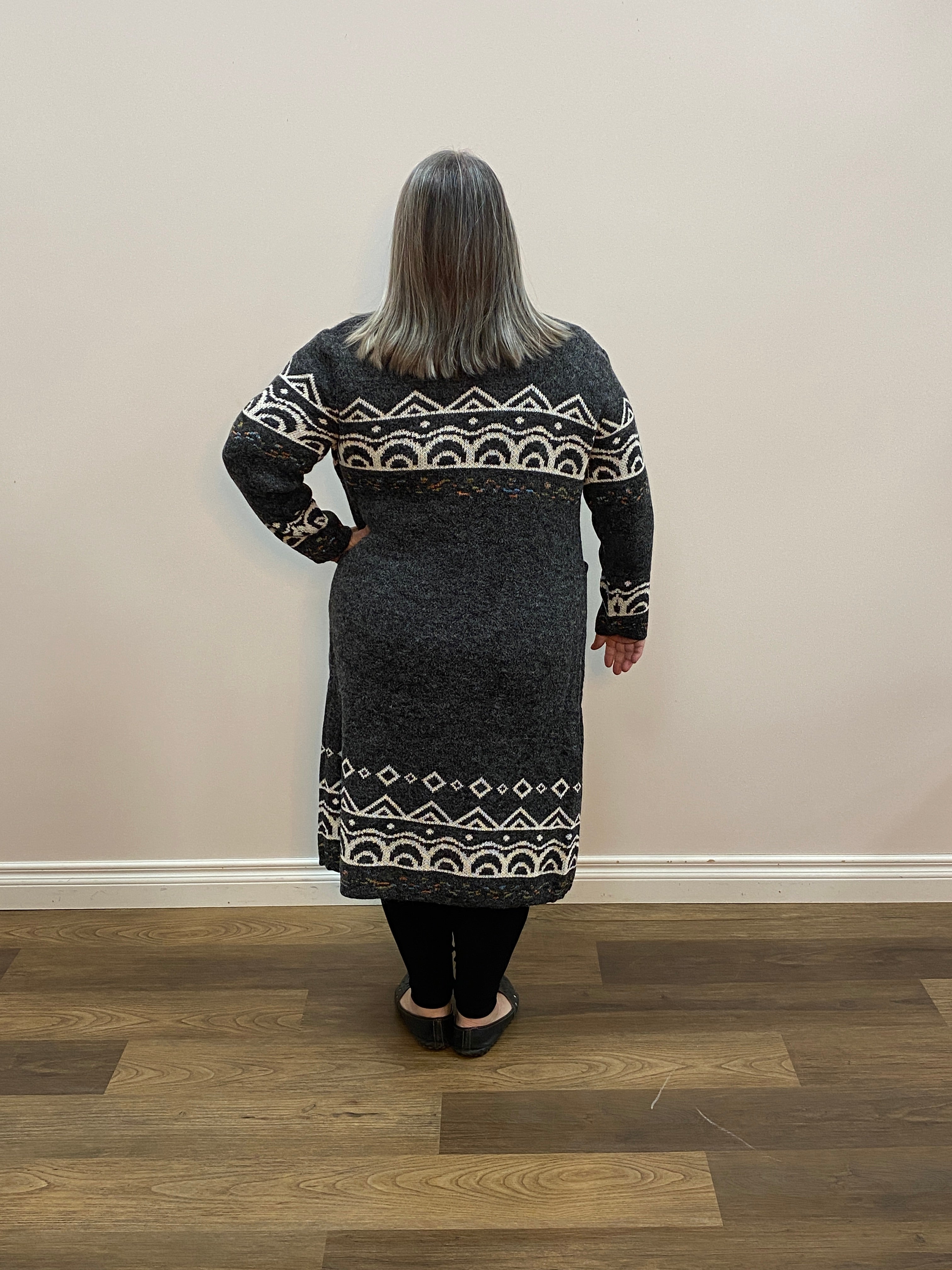 Person wearing a dark gray sweater with white patterns standing in a room with light-colored walls and wooden flooring.