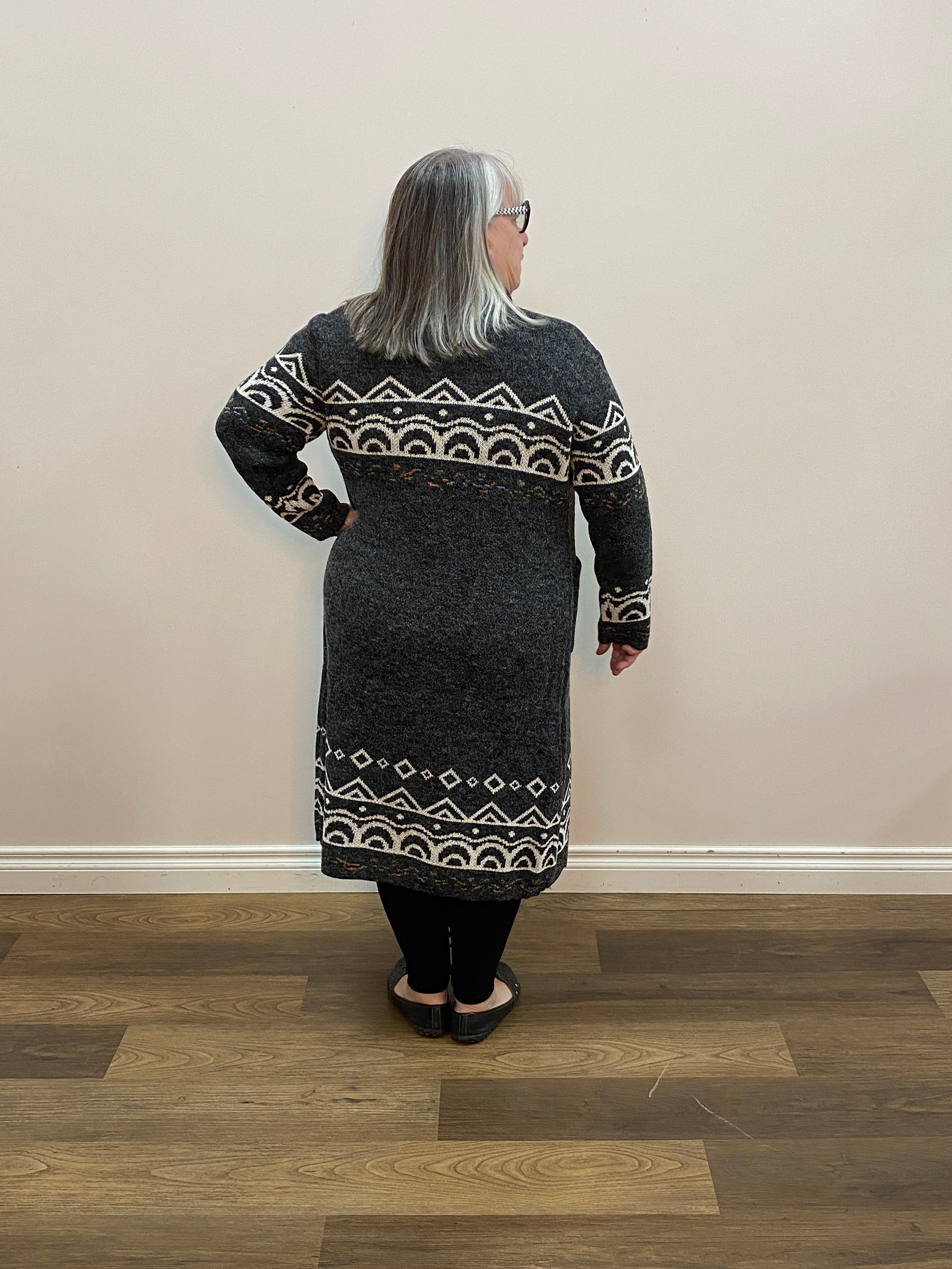 Person wearing a dark gray sweater with white patterns standing in a room with wooden flooring.