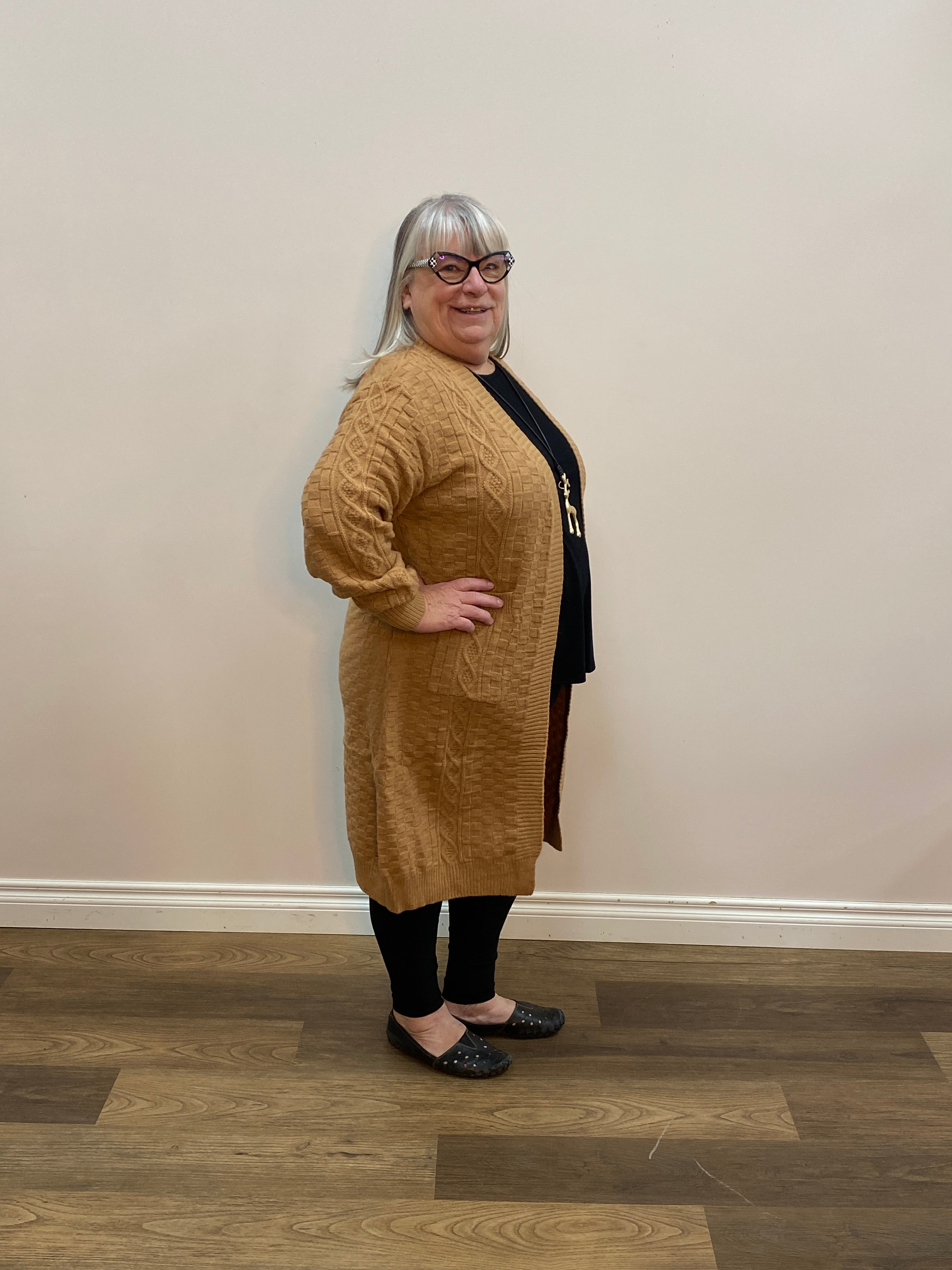 Person wearing a brown knitted cardigan in a room with wooden flooring and a white wall.