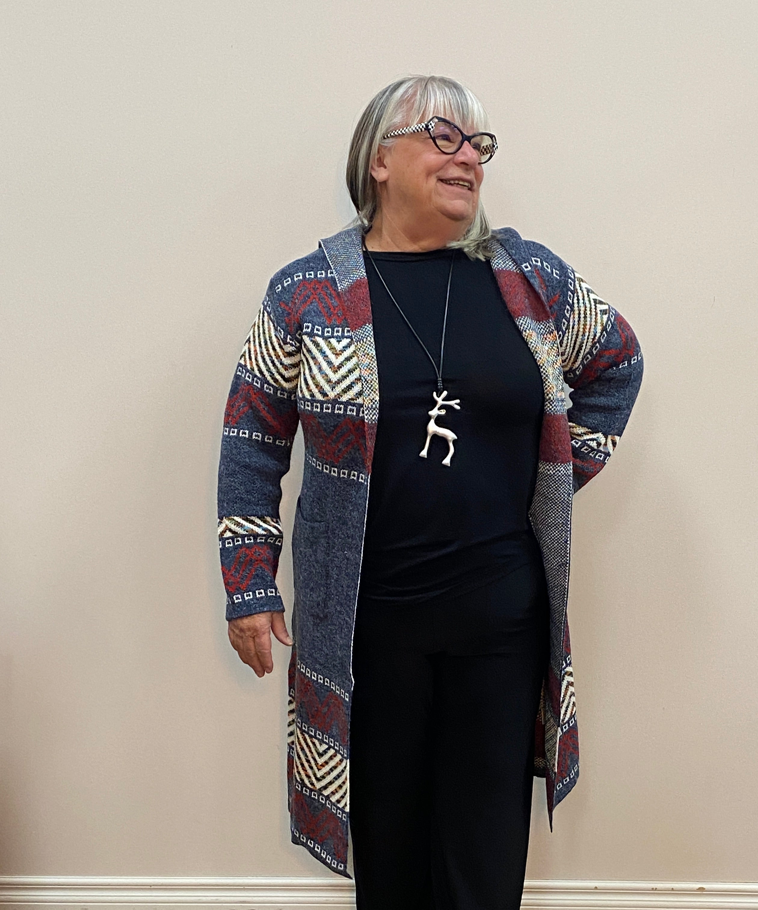 Person wearing a patterned jacket and black pants standing against a plain wall.