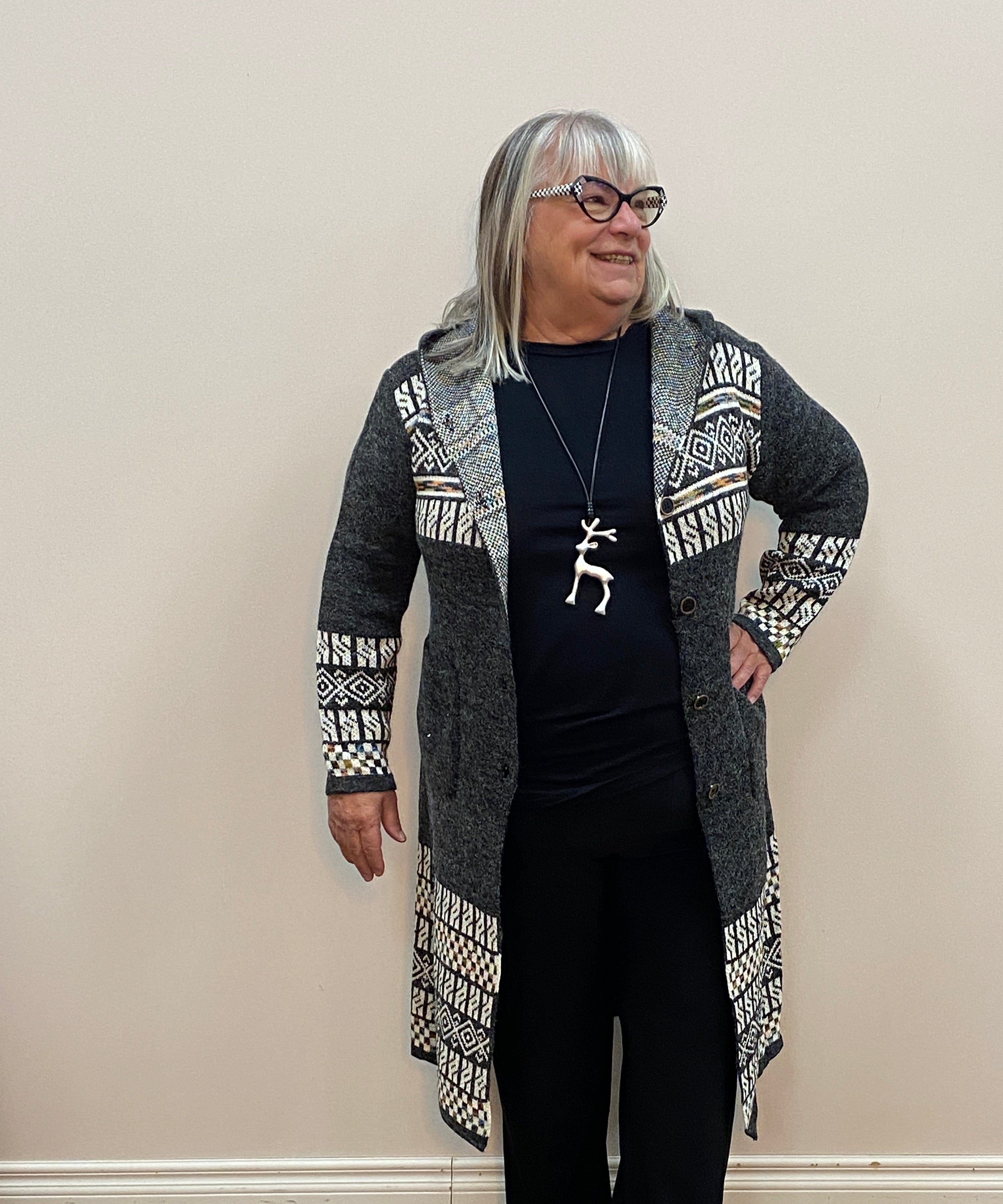 Person wearing a patterned cardigan and black pants standing against a white wall.