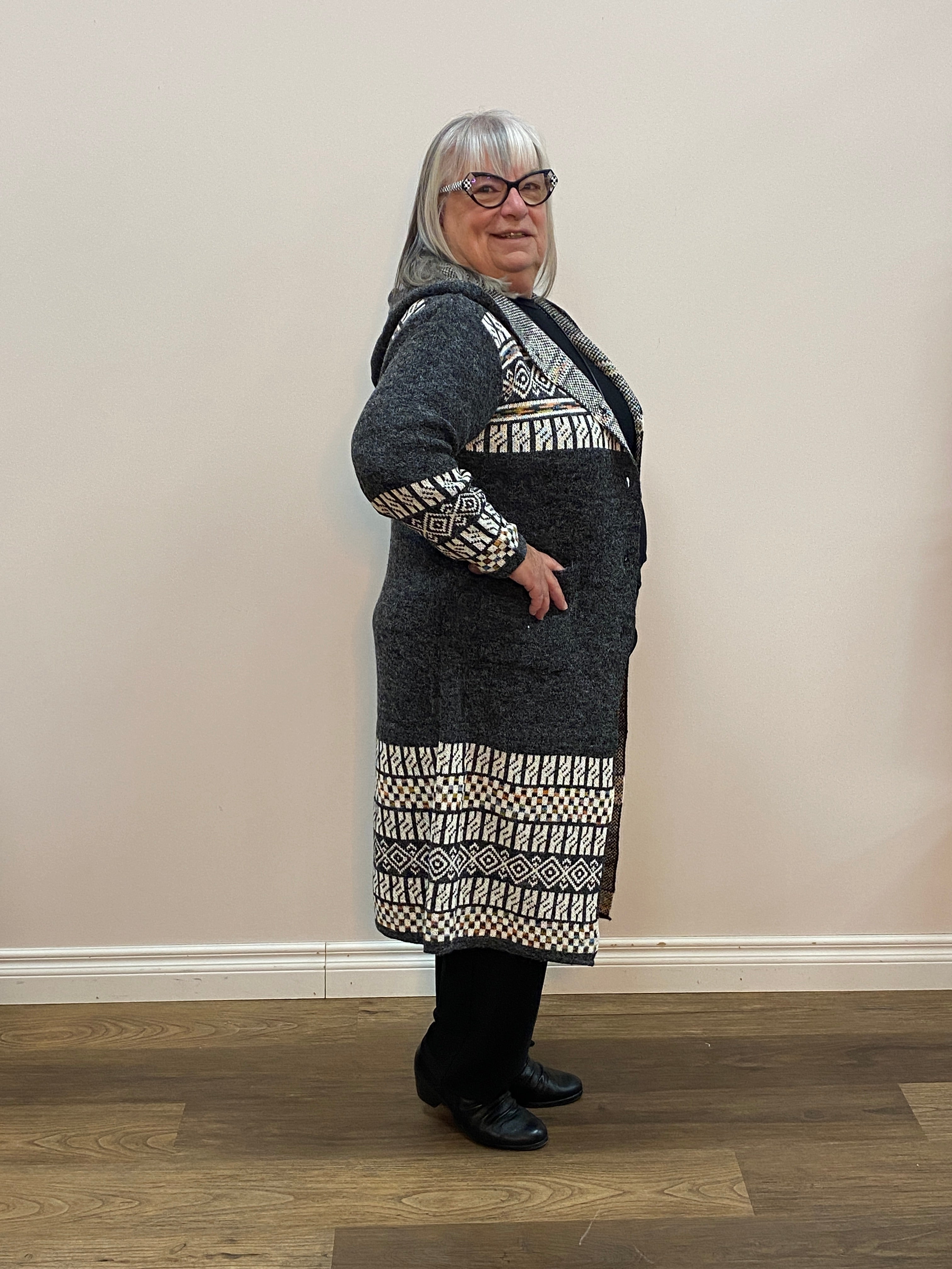 Person wearing a dark gray coat with white patterned sleeves standing against a plain wall.