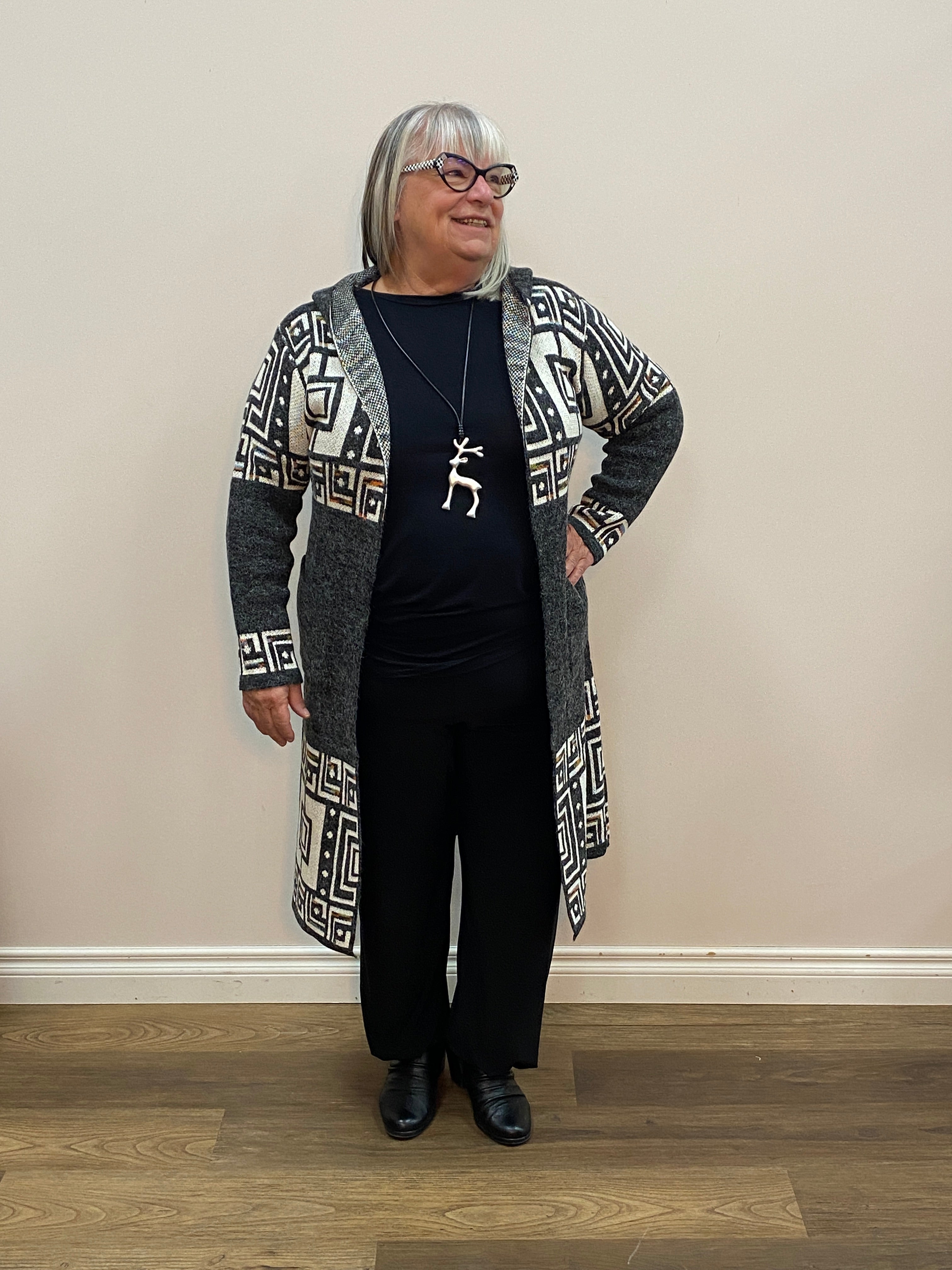 Person wearing a patterned coat and black pants standing against a plain wall.