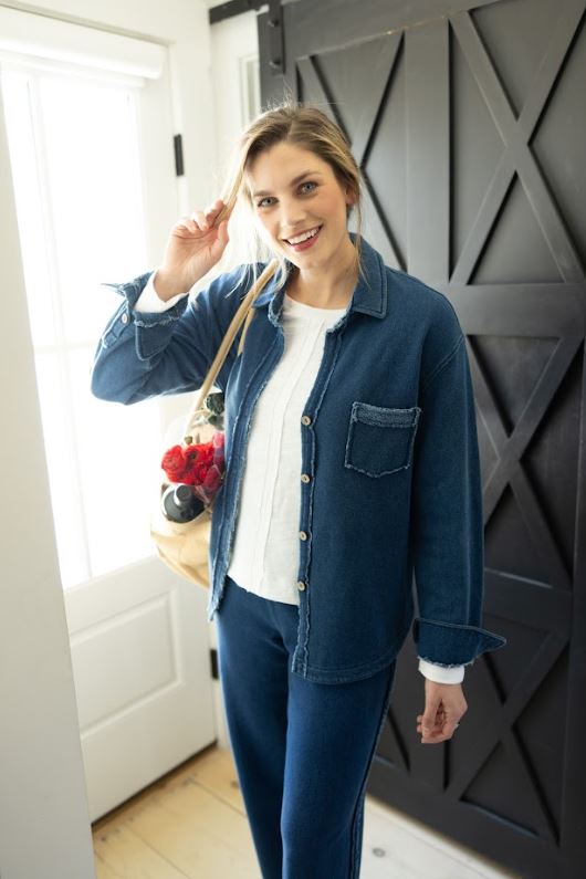 Woman in a blue denim shirt and pants standing indoors with a decorative wall in the background.