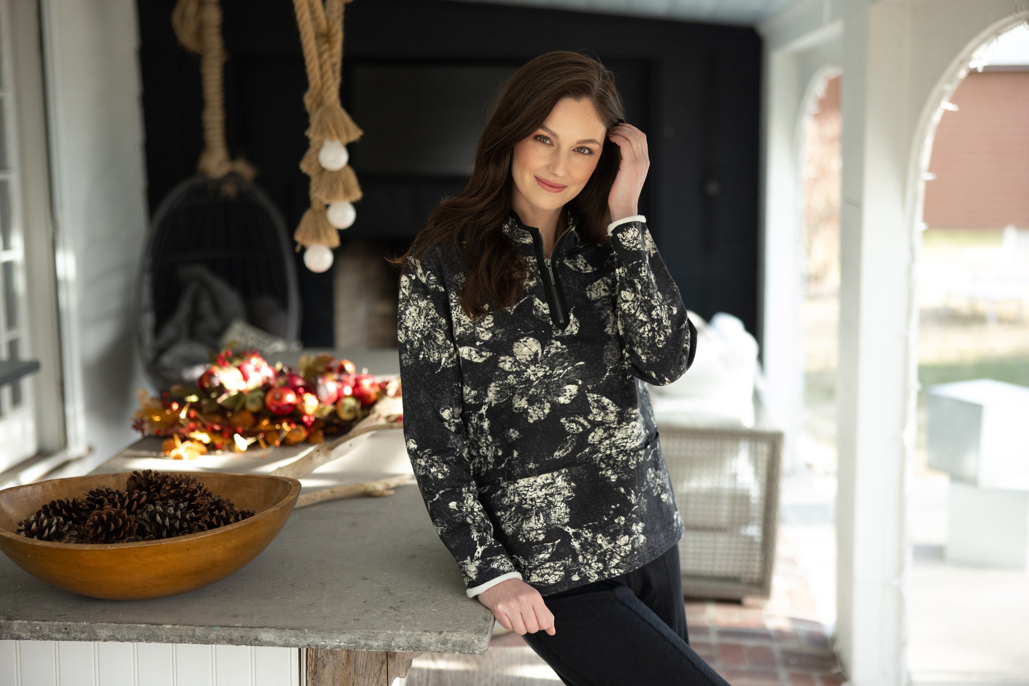 Woman in a floral patterned jacket standing in a room with a bowl of fruit and a fireplace.
