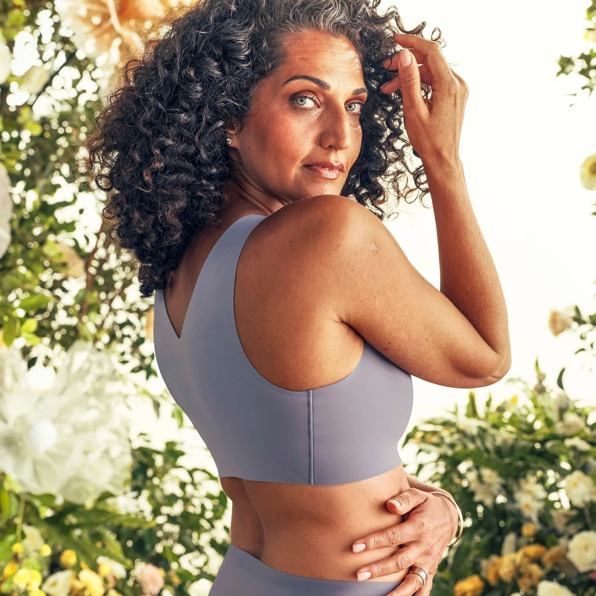 Woman wearing the Evelyn Bobbie Defy Bra in London Fog, light grey no wire bra, with a greenery background showing the back and side of bra.