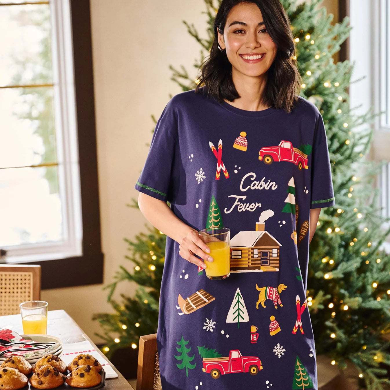 Woman wearing a 'Cabin Fever' themed nightshirt in a festive room with Christmas trees.