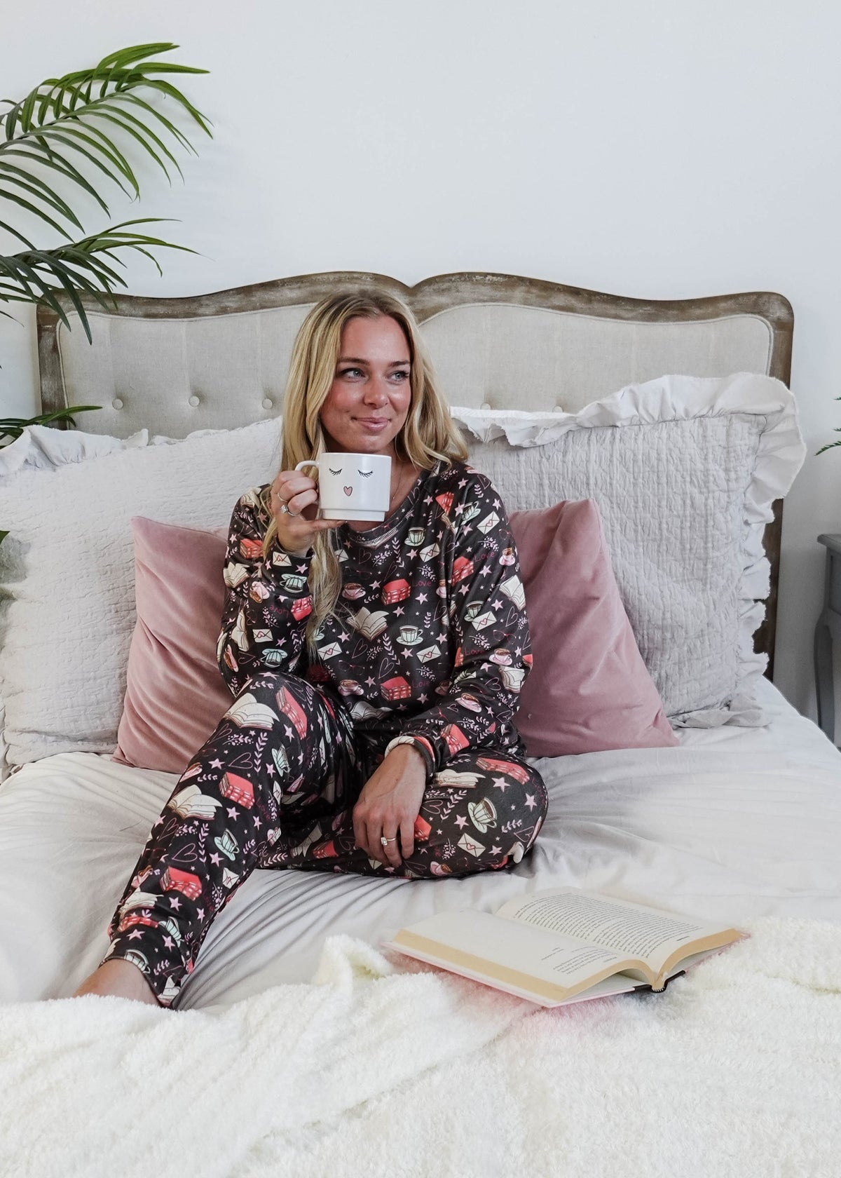 Woman in pajamas holding a mug on a bed with a plant in the corner