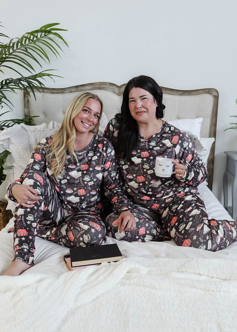 Two women in matching pajamas sitting on a bed with a plant in the background