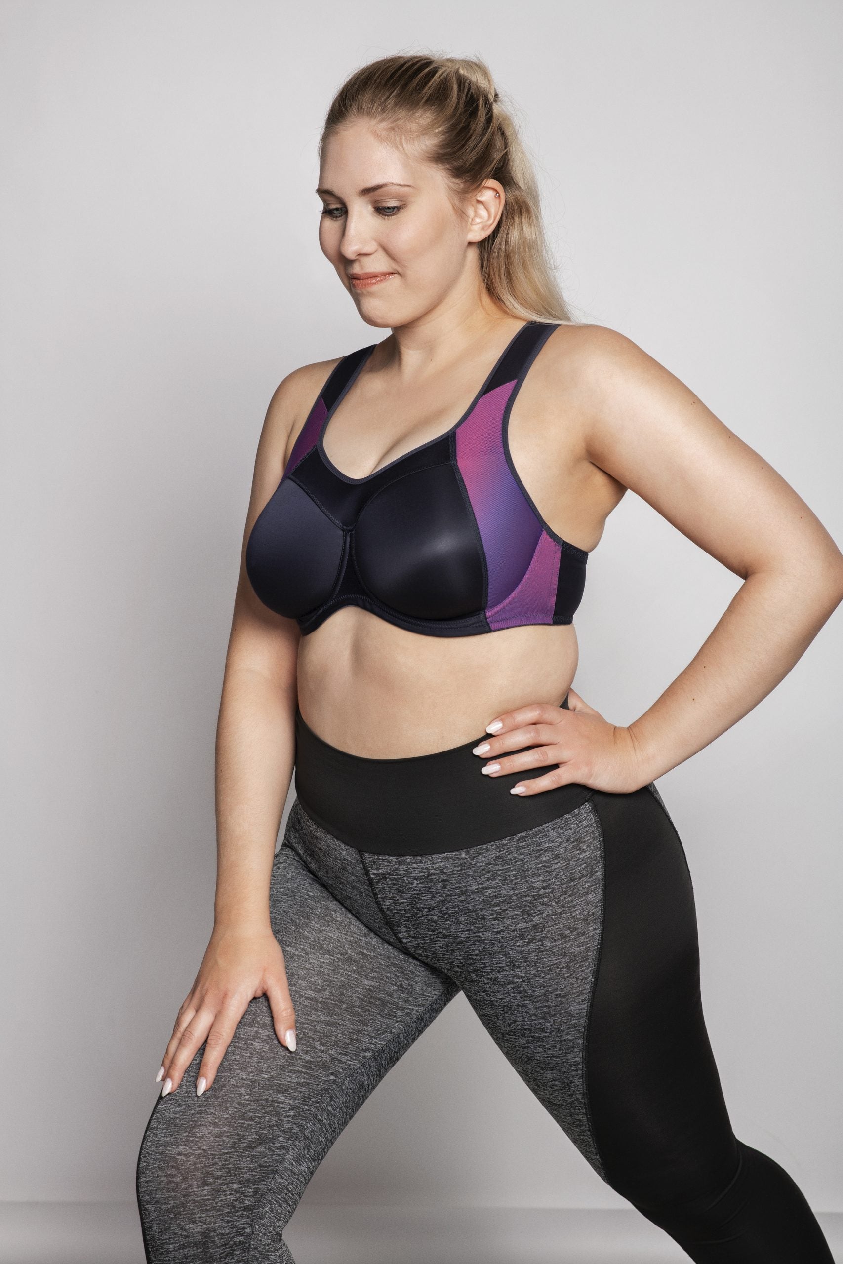 Woman wearing a black and purple sports bra and gray leggings on a plain background