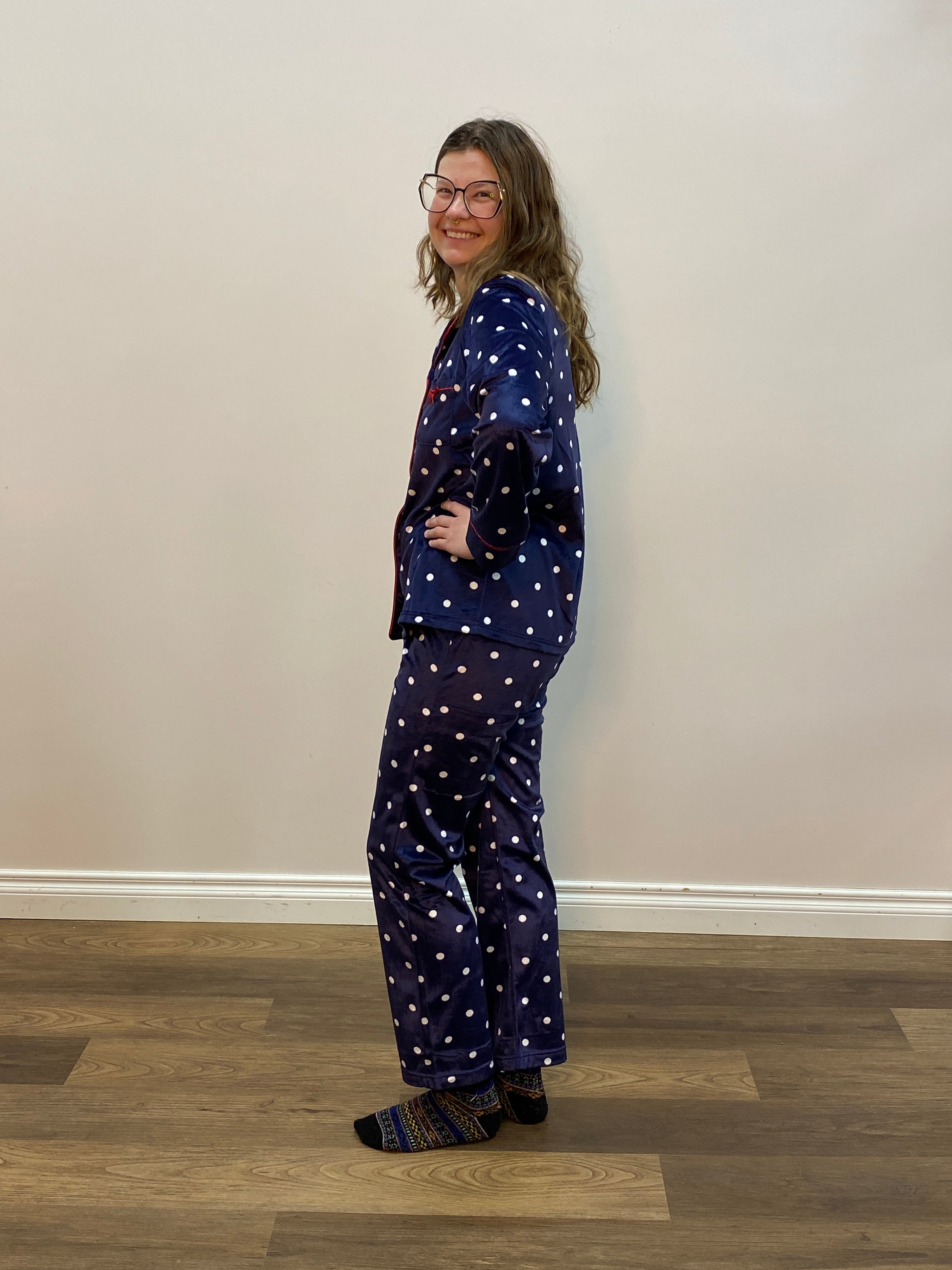Person wearing a navy blue polka pajama set standing in a room with a white wall and wooden floor.