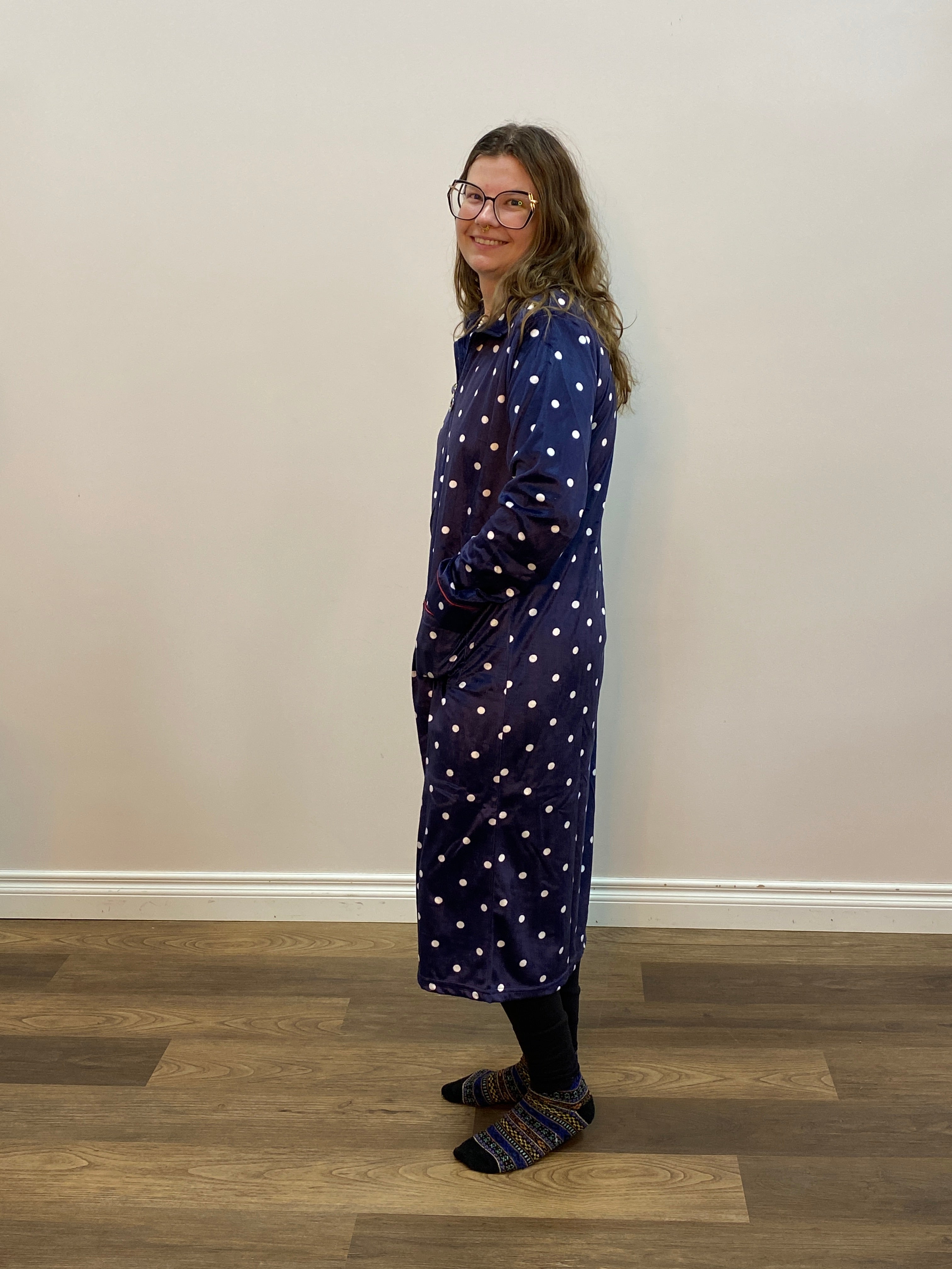 Person wearing a navy blue polka dot robe standing in a room with a white wall and wooden floor.