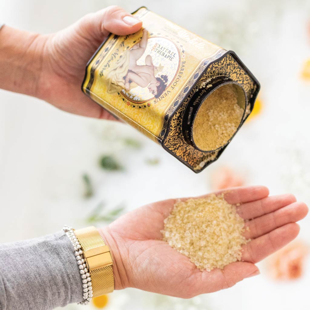 Barefoot Venus Mustard Bath in Yellow Tin container, shown against a white background, tipped on its side with some loose yellow bath salt poured into an outstretched hand.