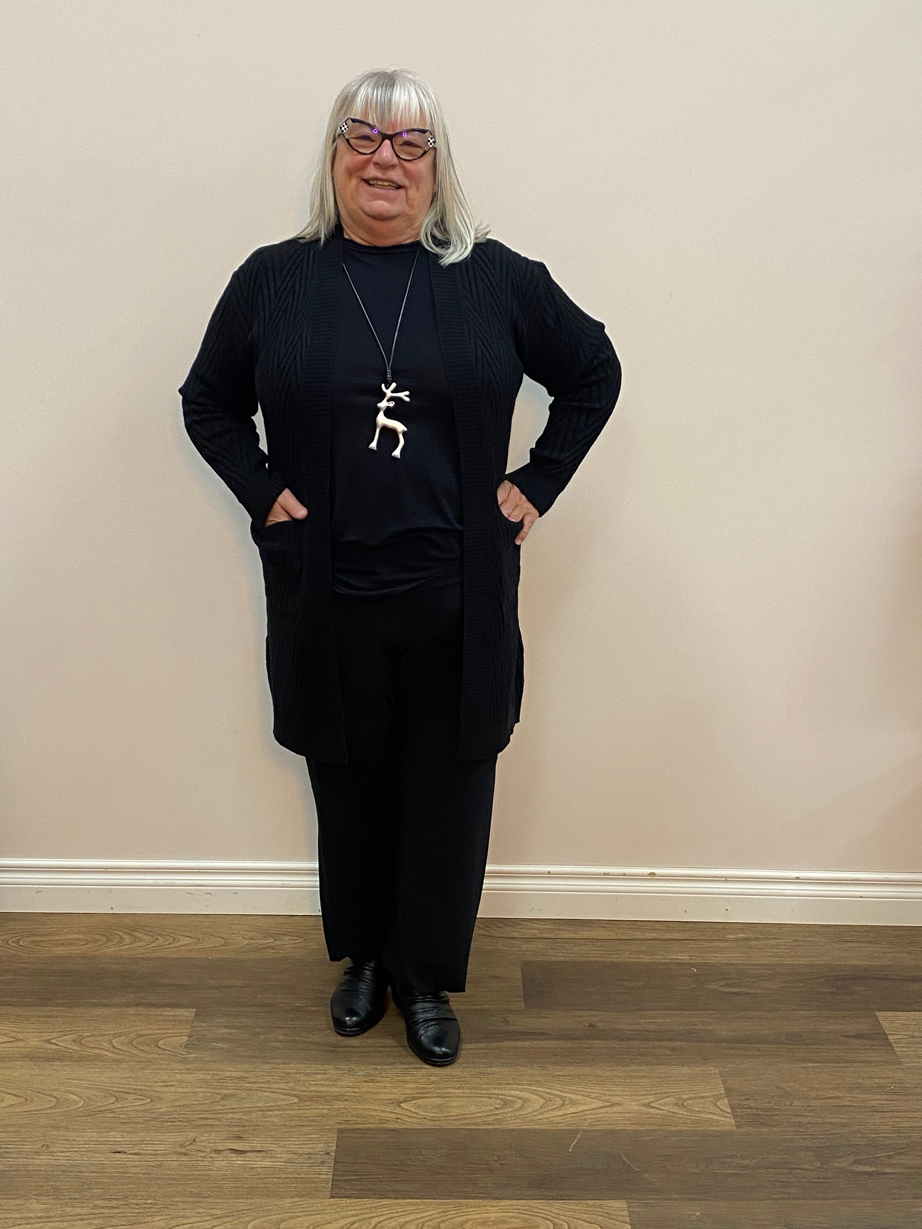 Person wearing a black outfit with a white necklace in a room with wooden flooring and a white wall.