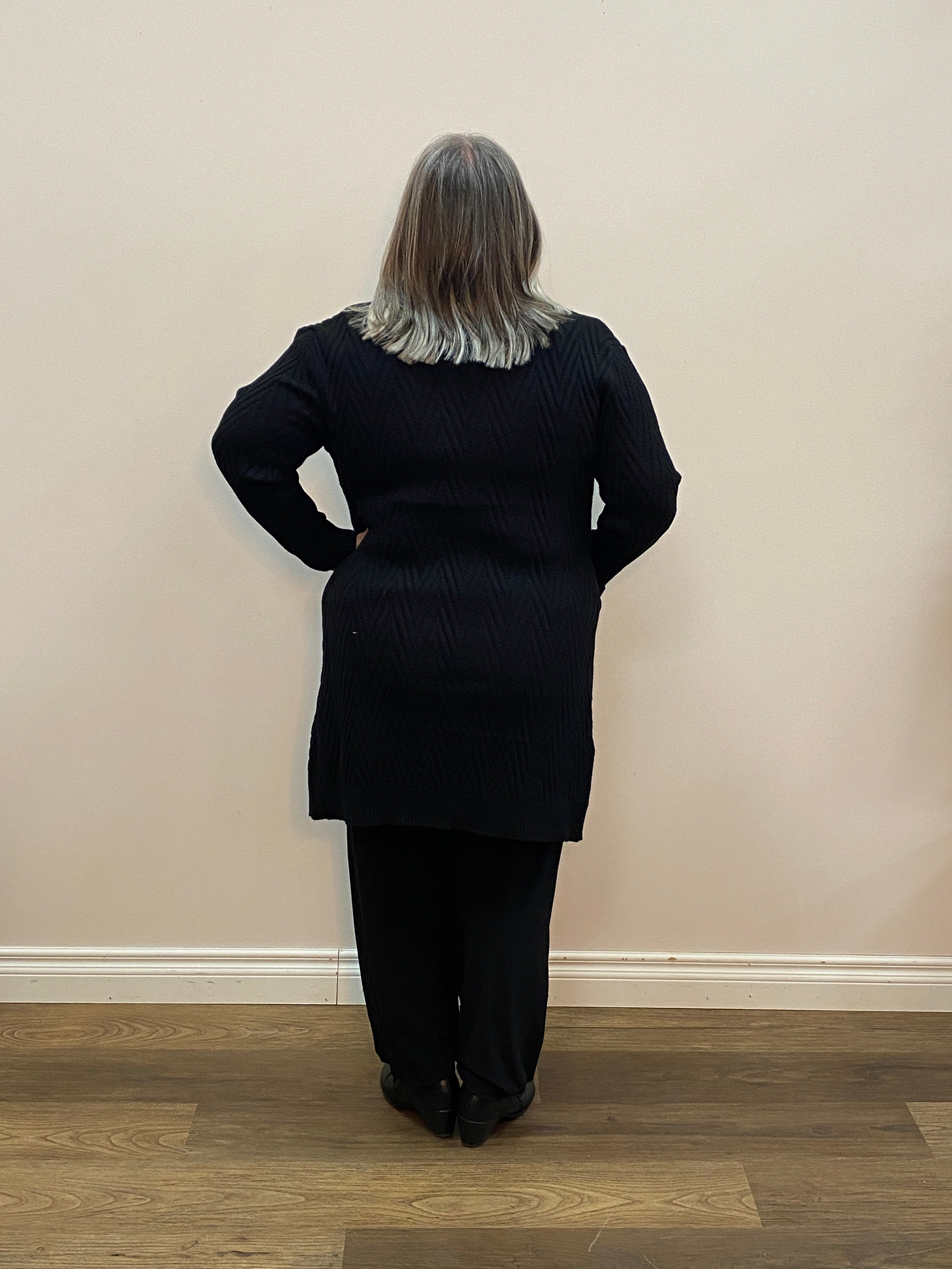Person wearing a black outfit standing against a plain wall.