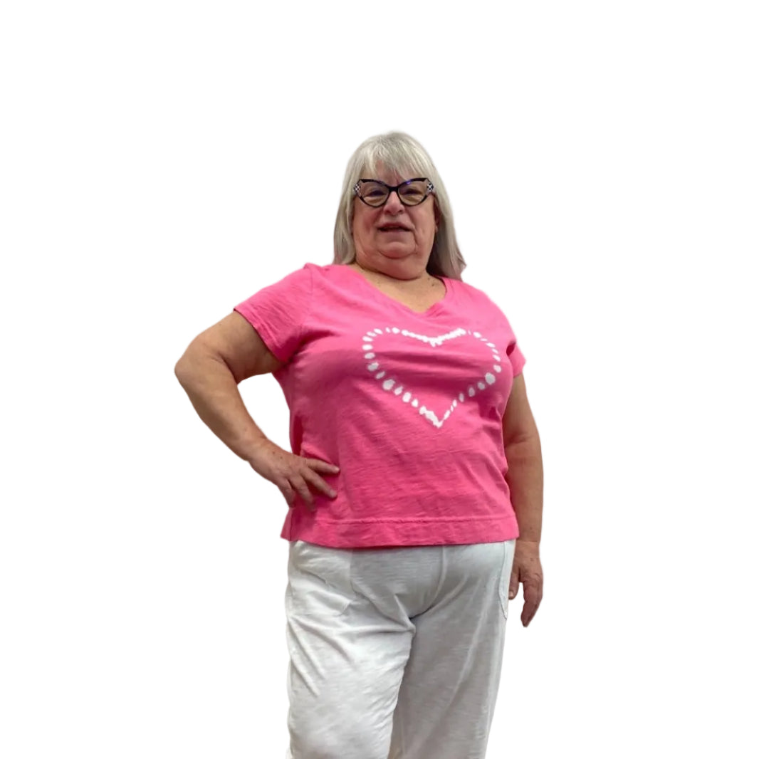 Woman wearing the Escape V Neck Tee in Cosmo Pink with a white heart shape on the front, paired with white pants, against a white background. 