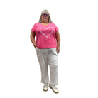 Woman wearing the Escape V Neck Tee in Cosmo Pink with a white heart shape on the front, paired with white pants, against a white background. 