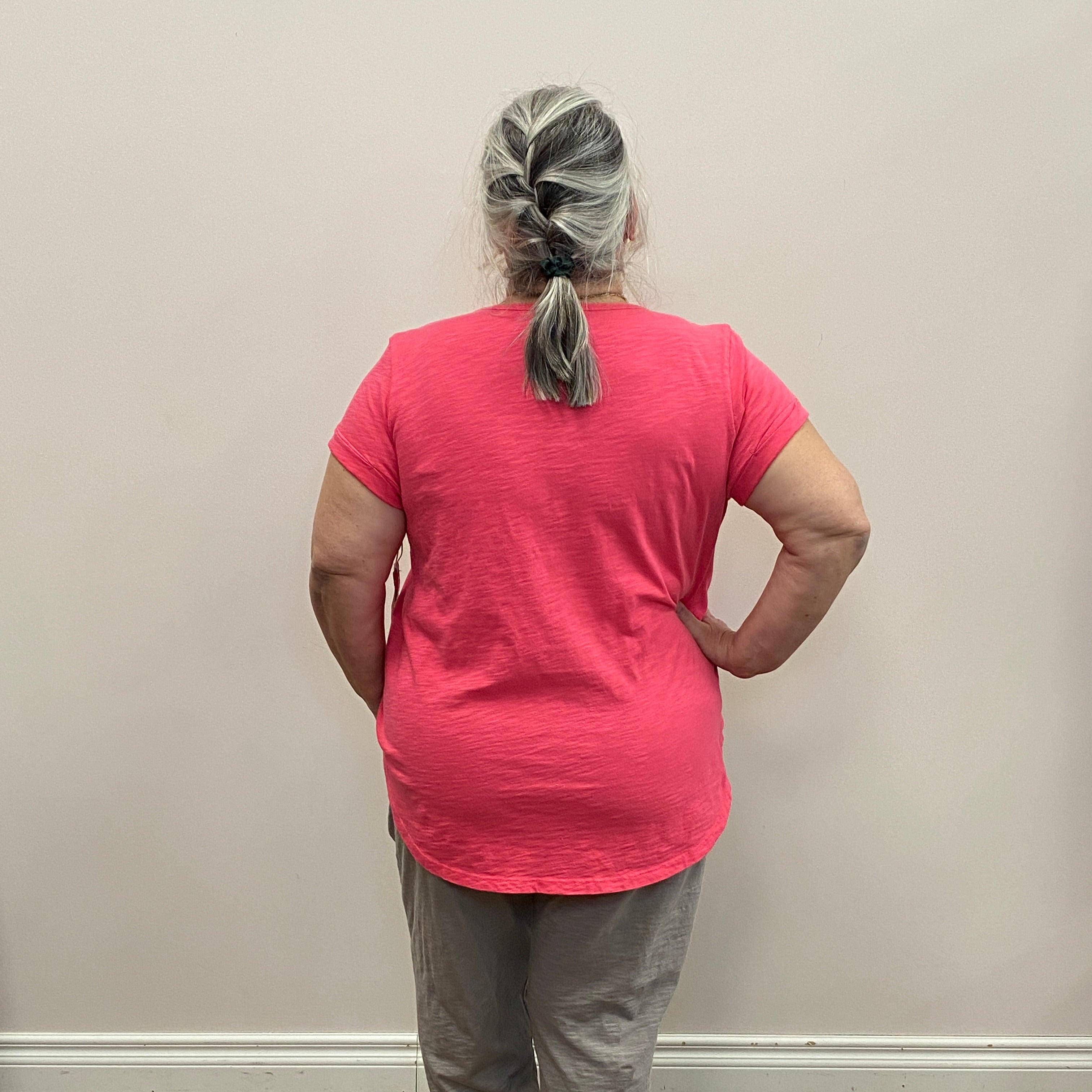 Person wearing a pink shirt and gray pants standing against a plain wall.