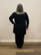 Person wearing a black coat standing in a room with wooden flooring and a white wall.