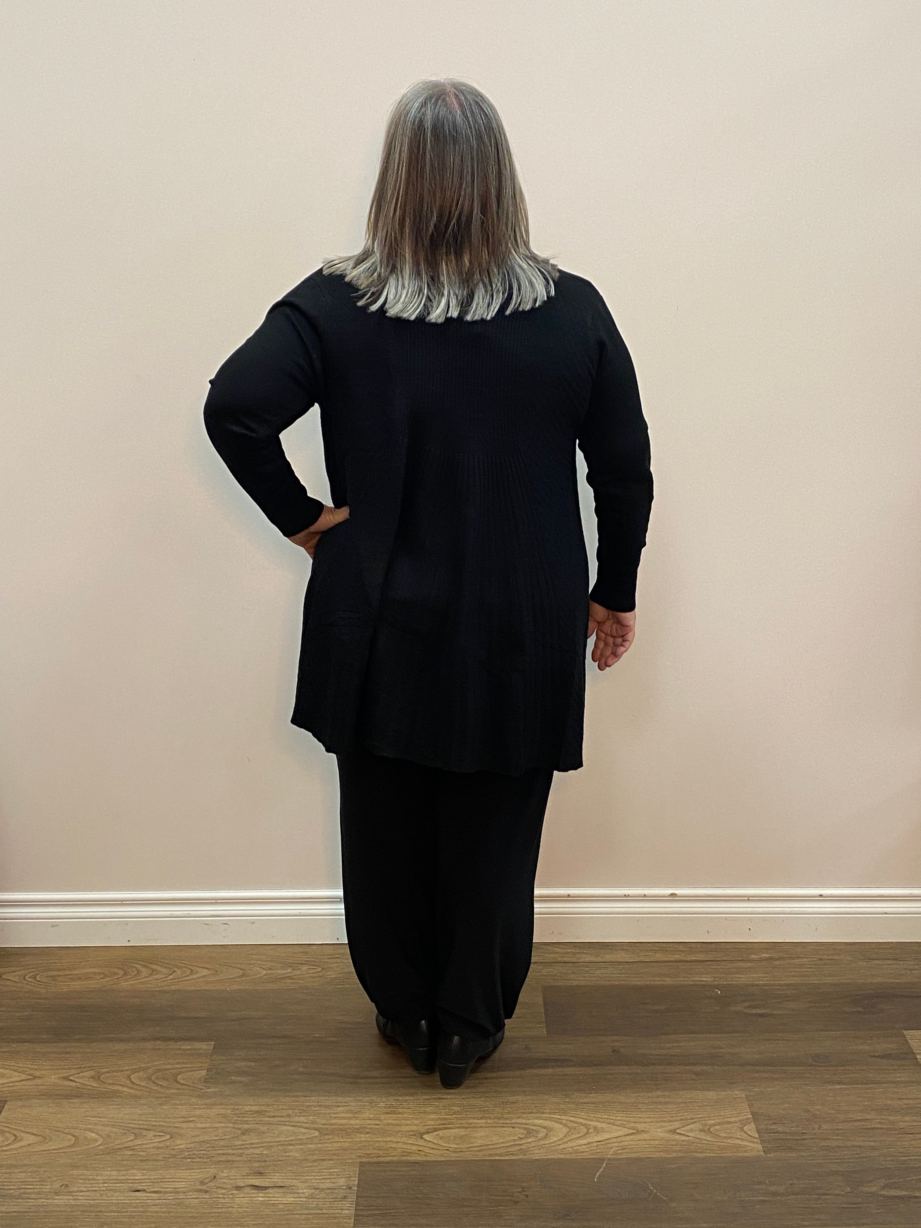 Person wearing a black coat standing in a room with wooden flooring and a white wall.