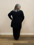Person wearing a black coat standing against a plain wall.