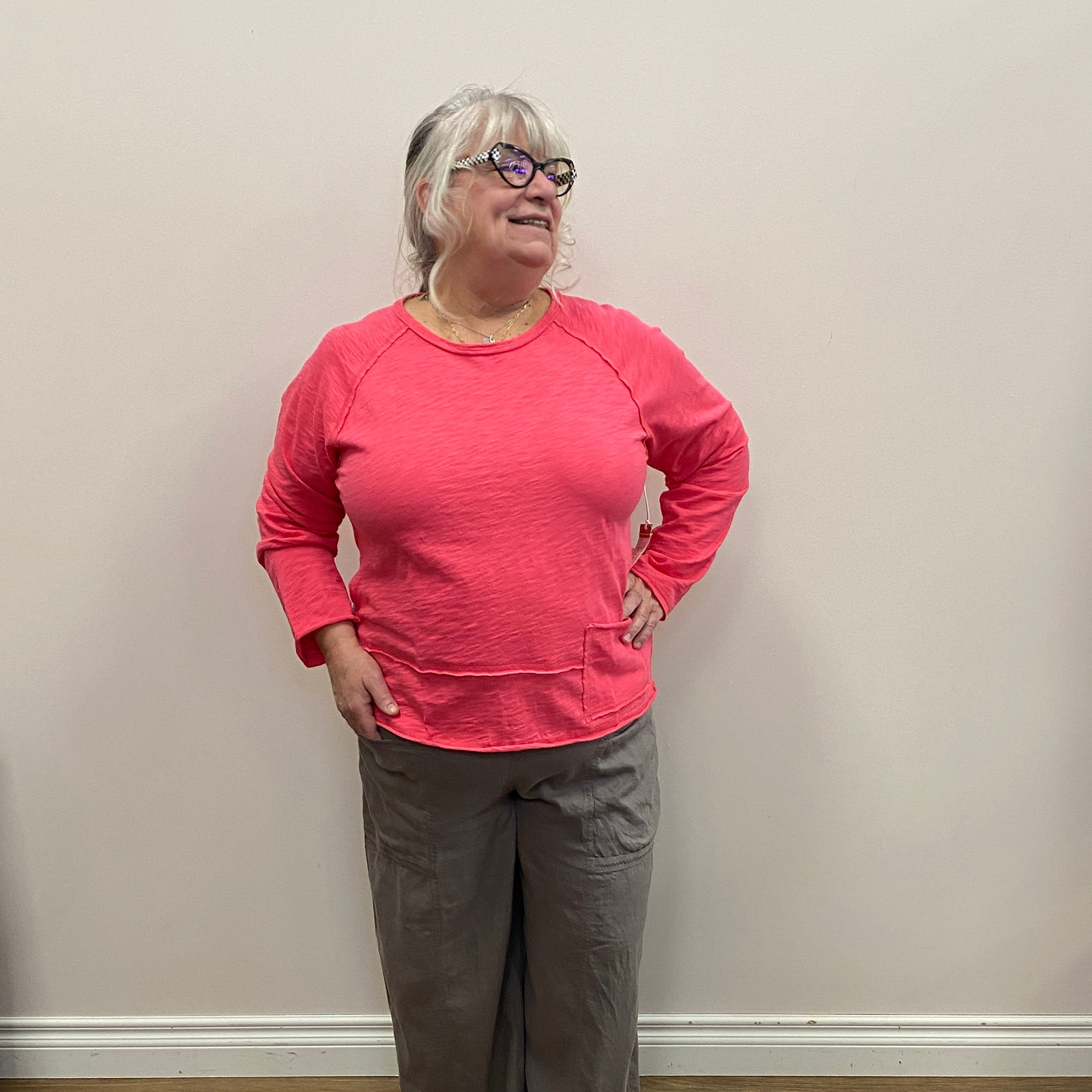 Person wearing a pink sweatshirt and dark pants standing against a white wall with a wooden floor.