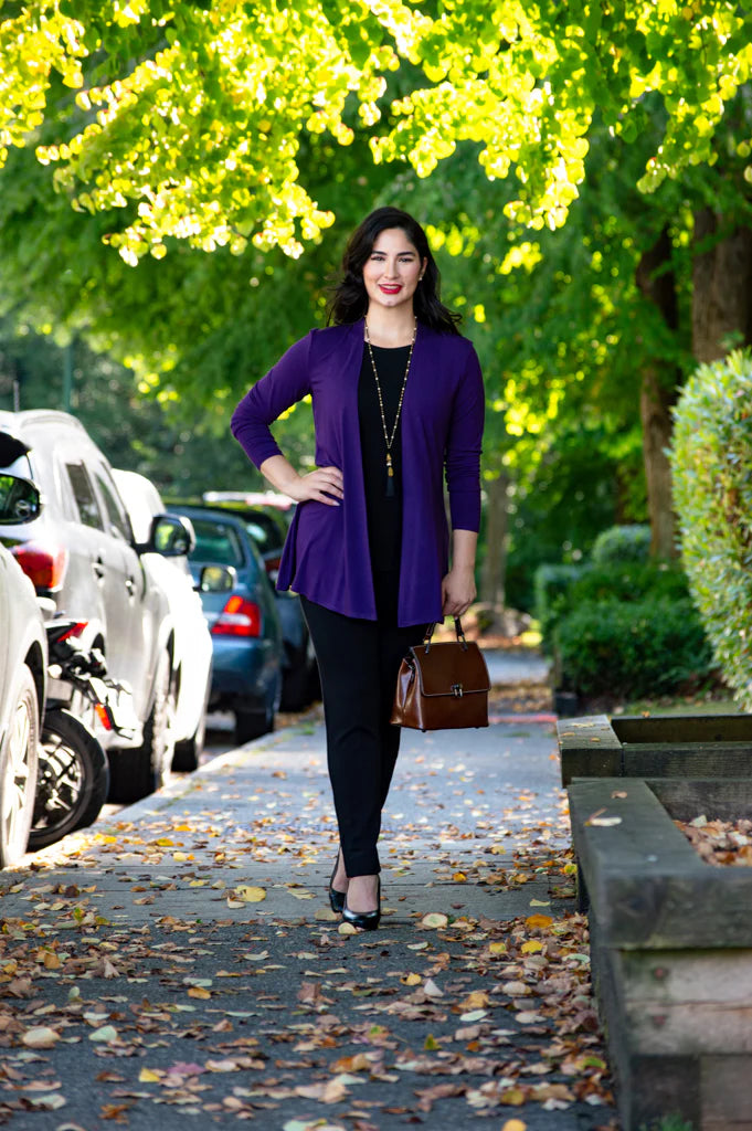 Woman in a purple cardigan walking on a sidewalk with cars and greenery in the background