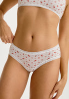The Blush Micro Hipster in Red Rose, white lace underwear with red floral pattern worn by a person on a plain background