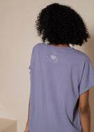 Person wearing a purple t-shirt with a small white logo on the back against a plain background