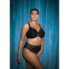 A woman wearing a black bra and panty set with lace details against a teal curtain. 