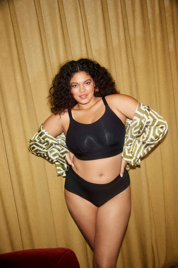 A woman wearing a black wireless bra and panty set against a gold curtain. 