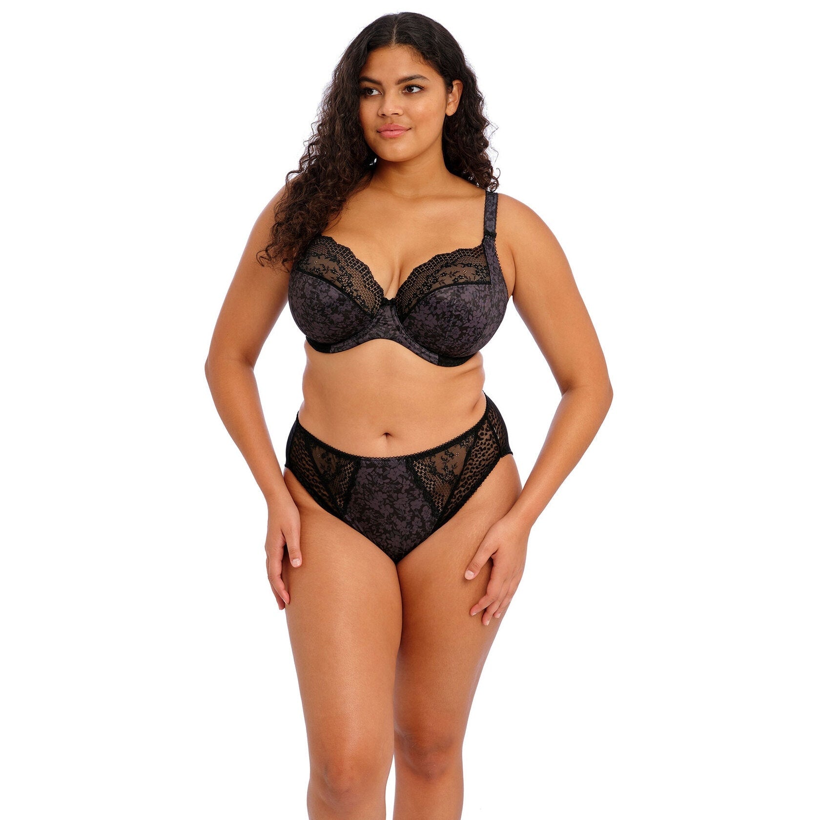 A woman wearing a black lace bra and high rise panty set against a white back ground. 