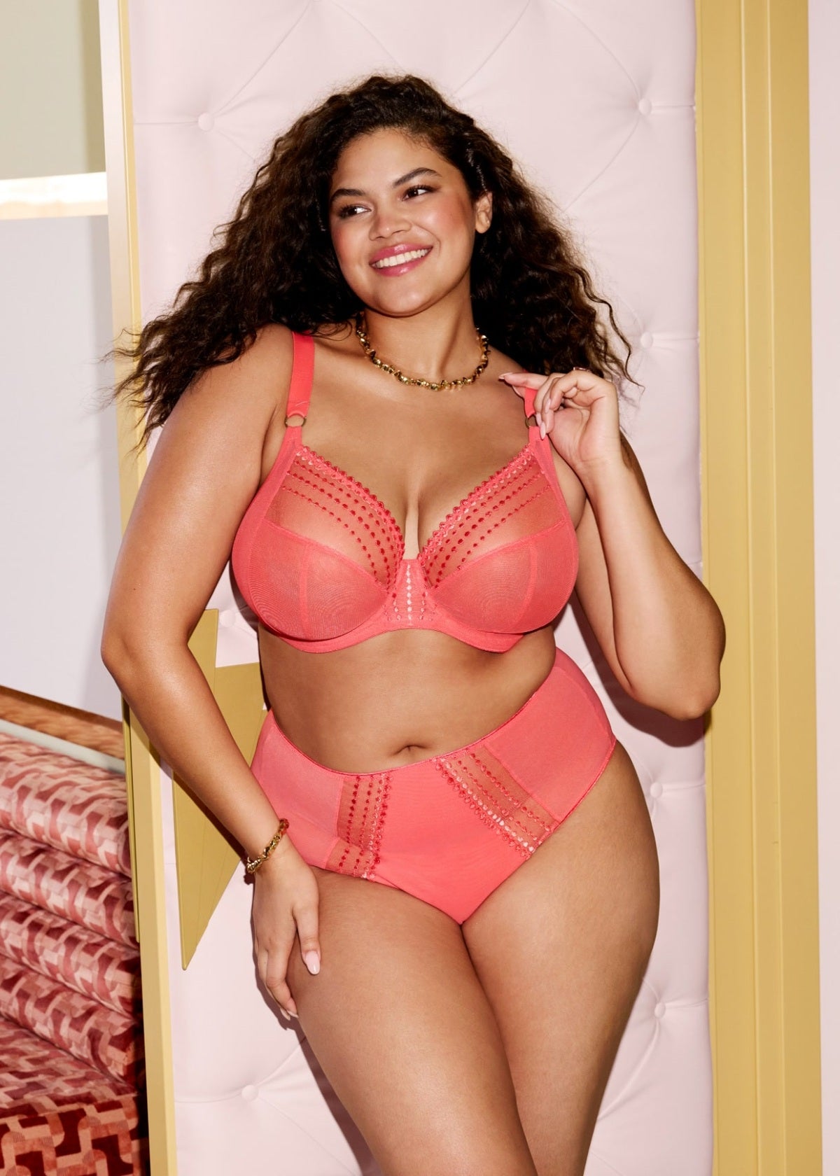 Woman wearing a coral bra and panties set in a bedroom setting.