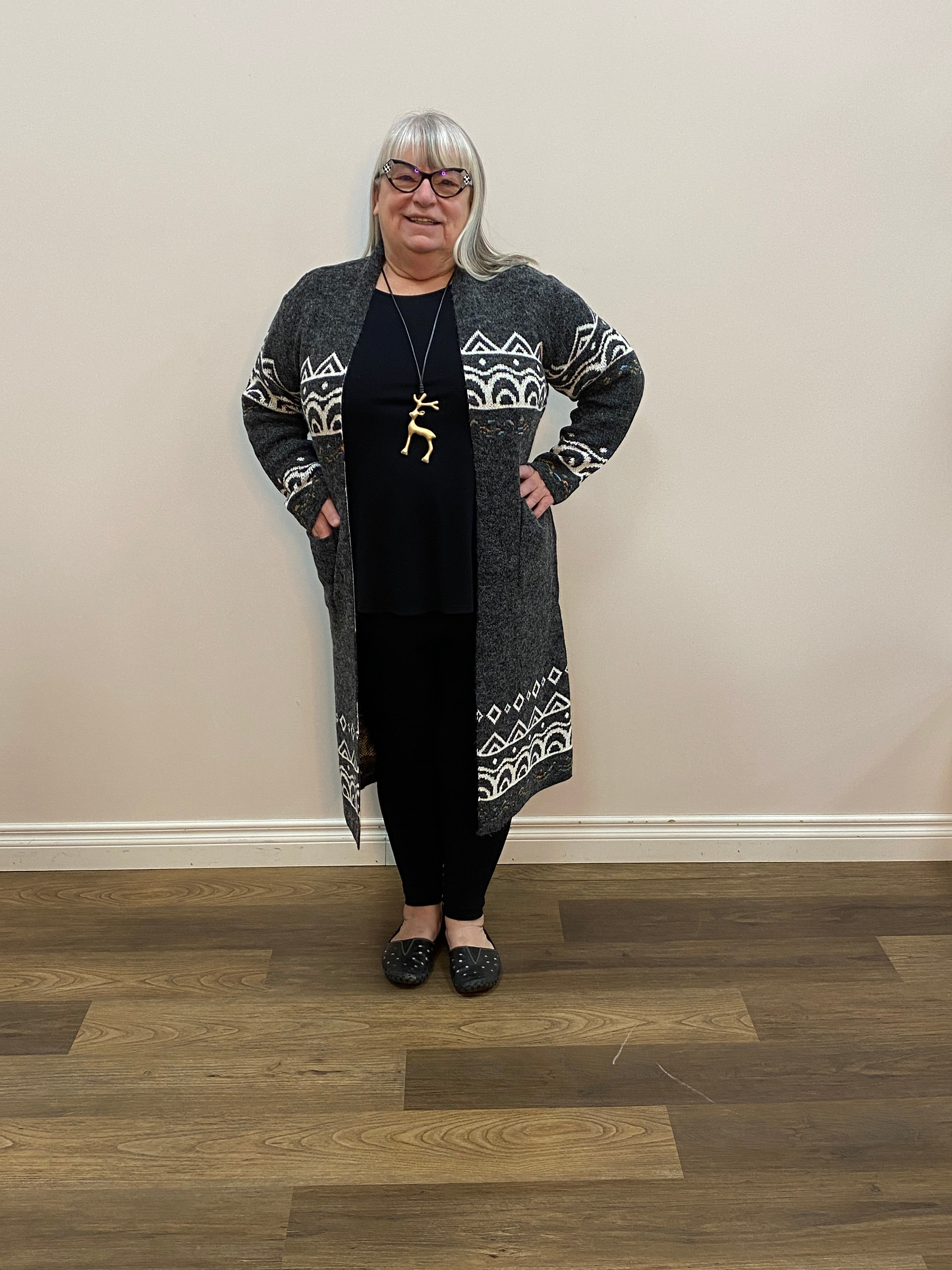 Person wearing a long dark cardigan with white patterns standing in a room with wooden flooring.
