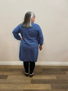Person wearing a blue textured sweater and black pants standing in a room with white walls and wooden flooring.
