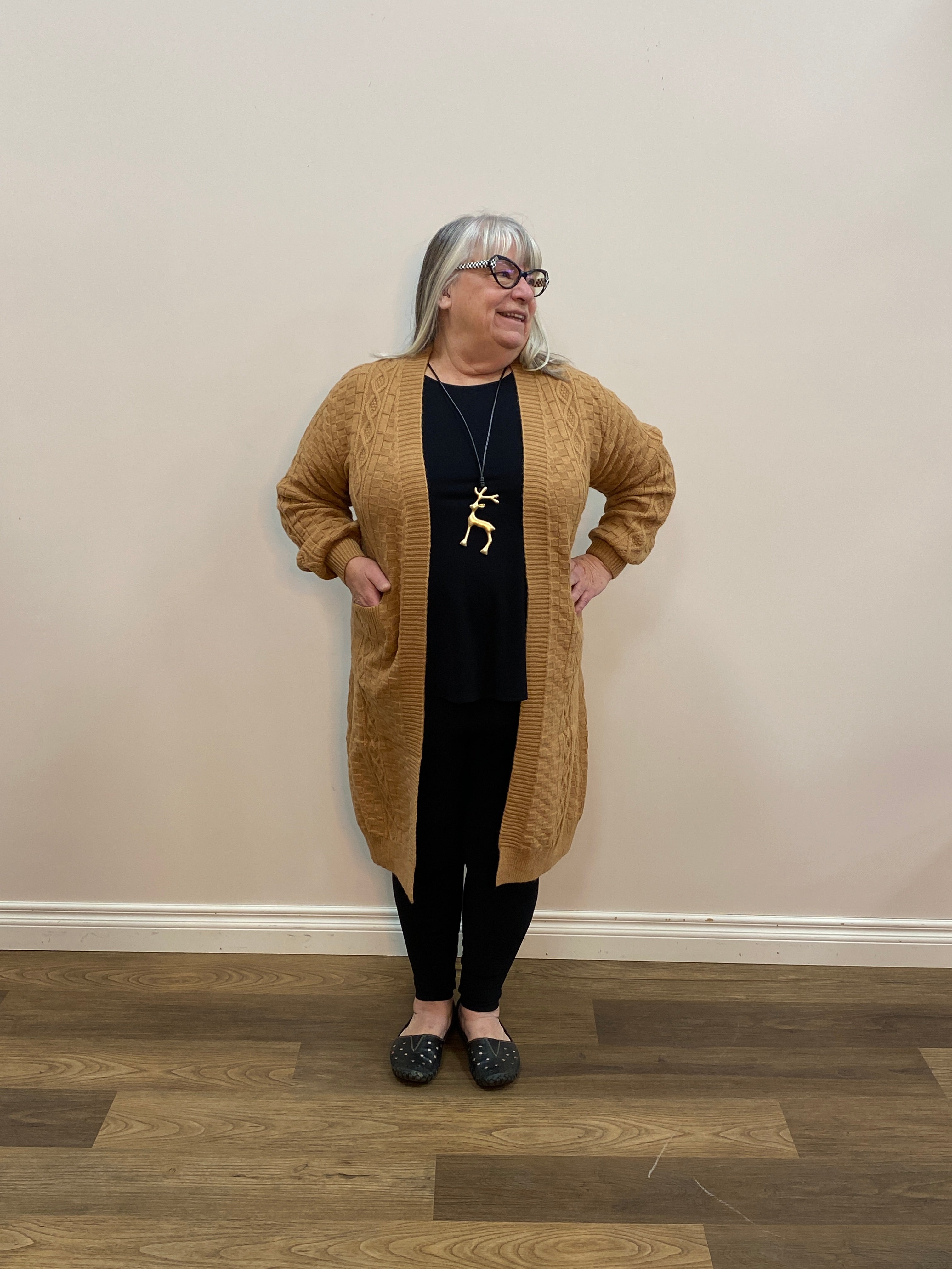 Person wearing a brown sweater and black pants standing in a room with wooden flooring.
