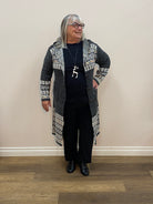 Person wearing a patterned cardigan and black pants standing against a white wall.