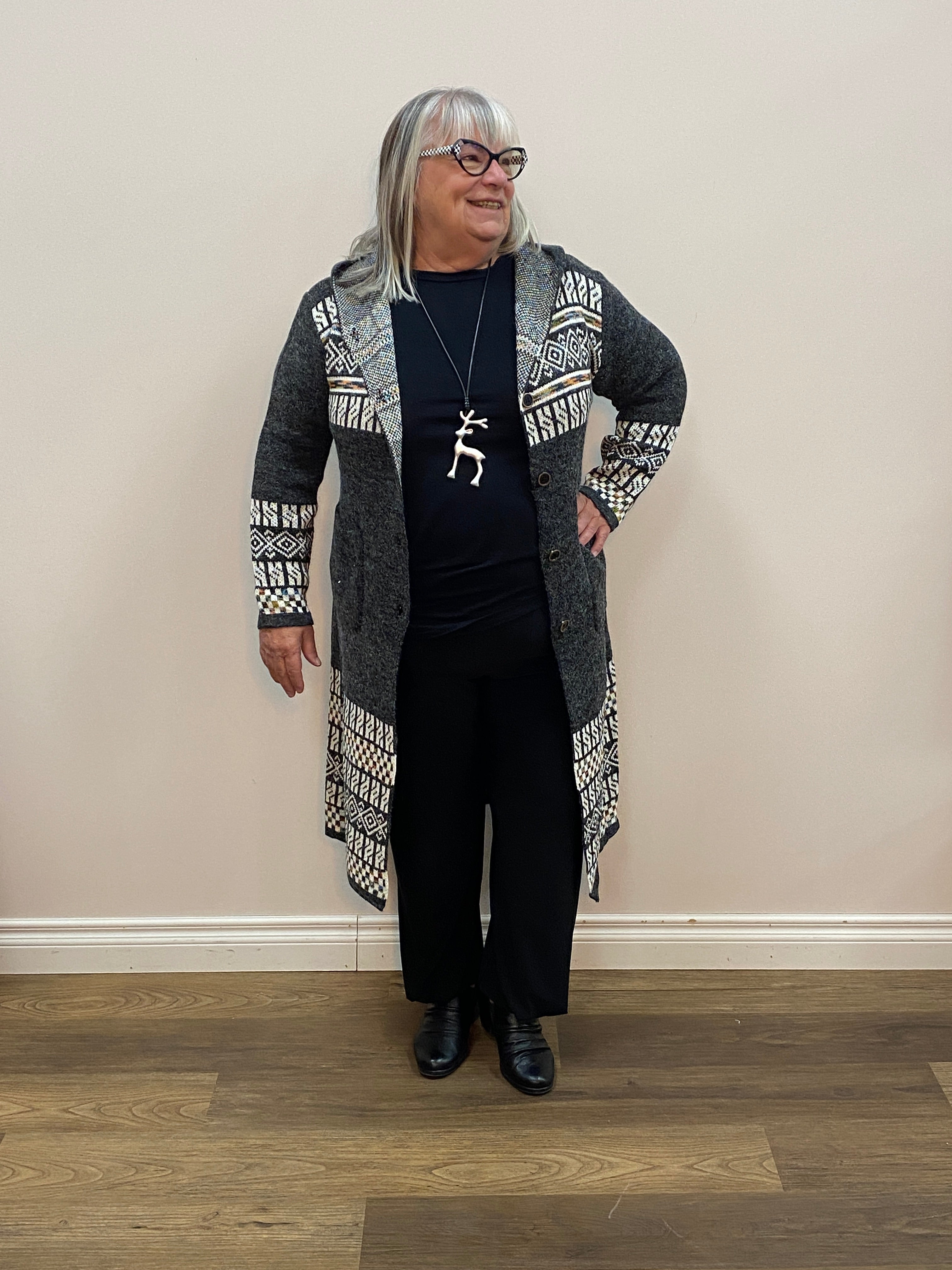Person wearing a patterned cardigan and black pants standing against a white wall.
