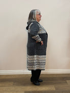 Person wearing a dark coat with patterned trim standing against a light-colored wall.