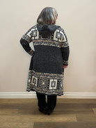 Person wearing a dark coat with geometric patterned lapels standing in a room with wooden floor and white walls.