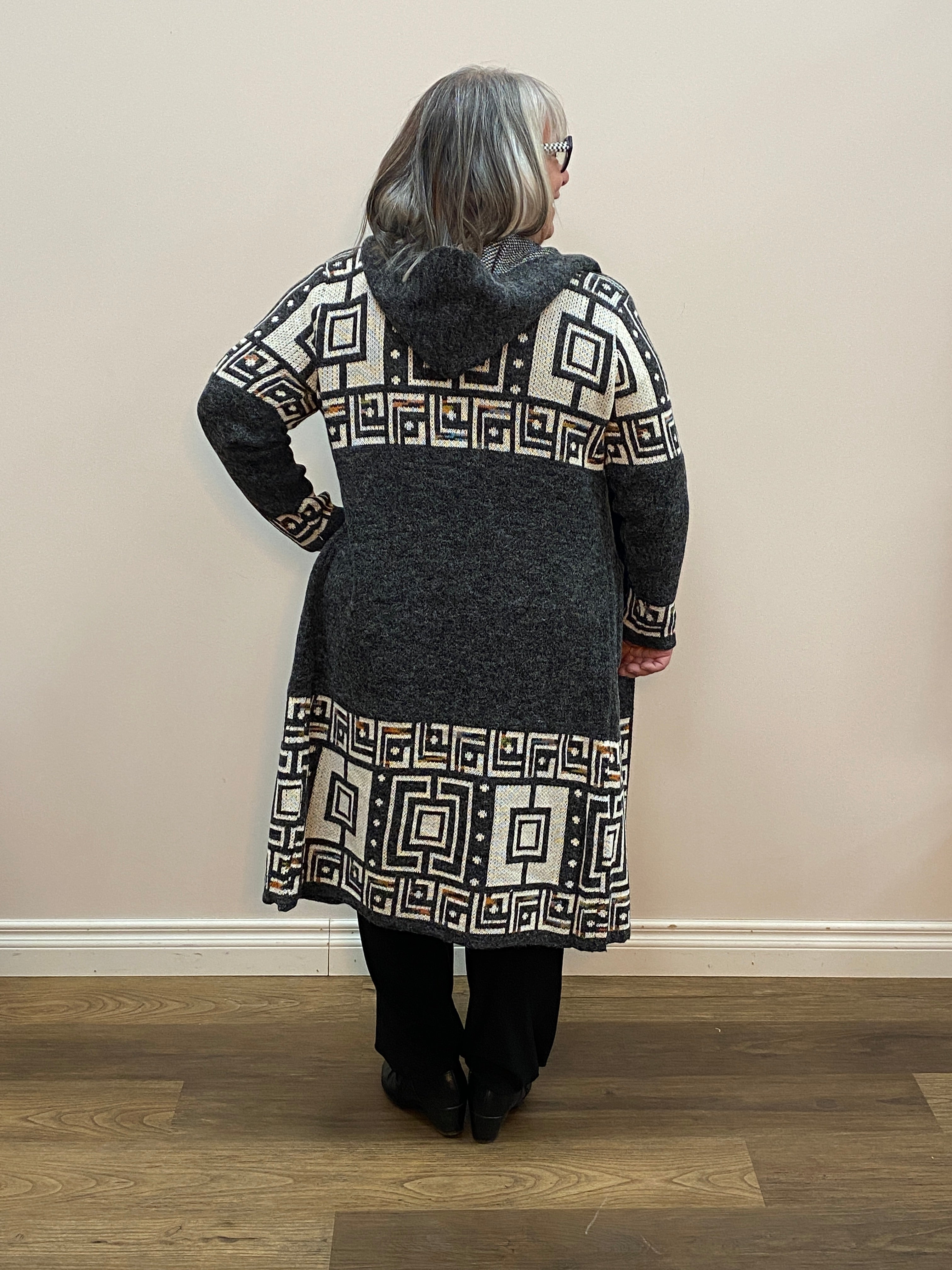 Person wearing a dark coat with geometric patterned lapels standing in a room with wooden floor and white walls.