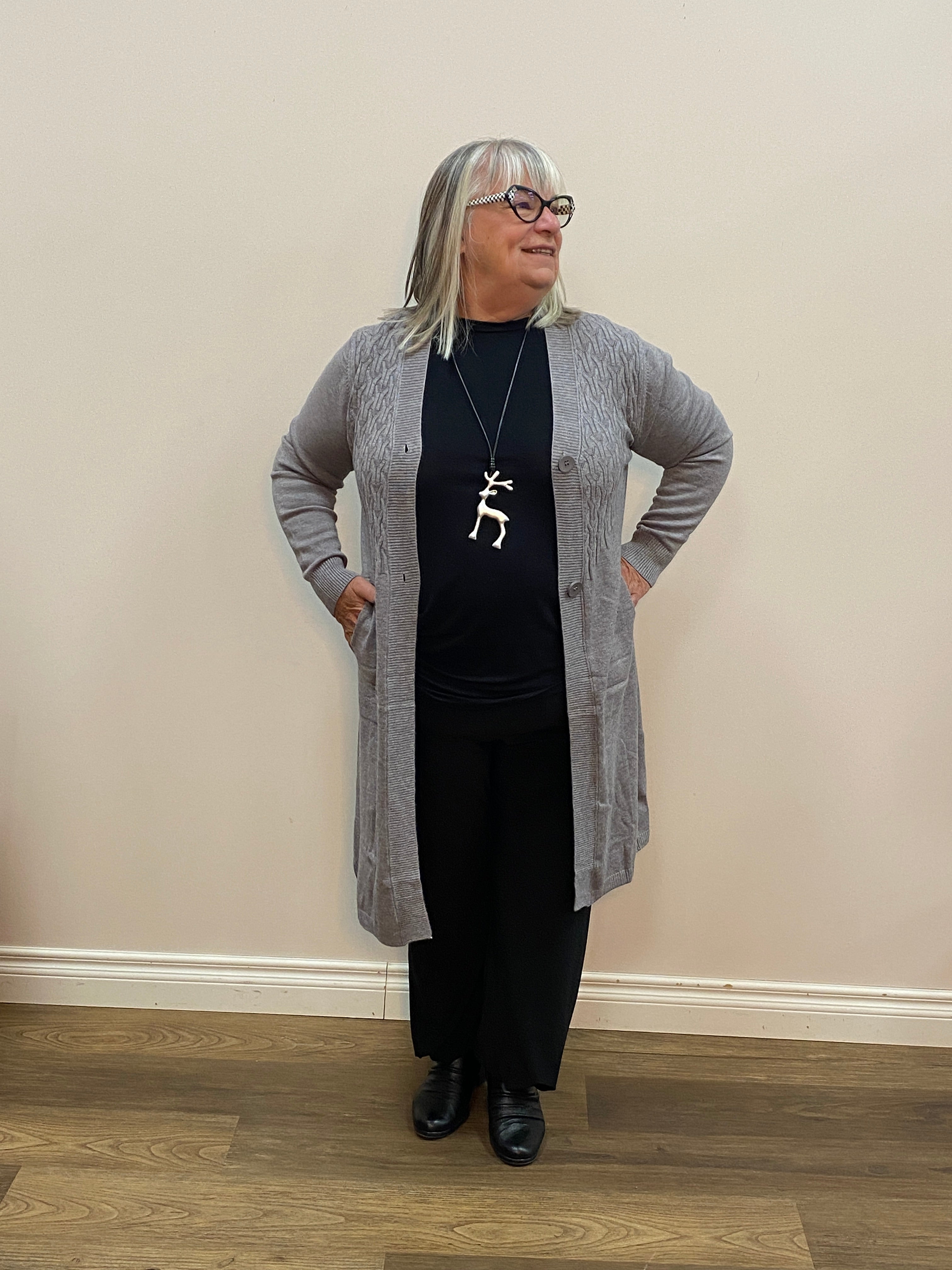 Person wearing a gray cardigan, black top, and black pants in a room with a wooden floor and white walls.