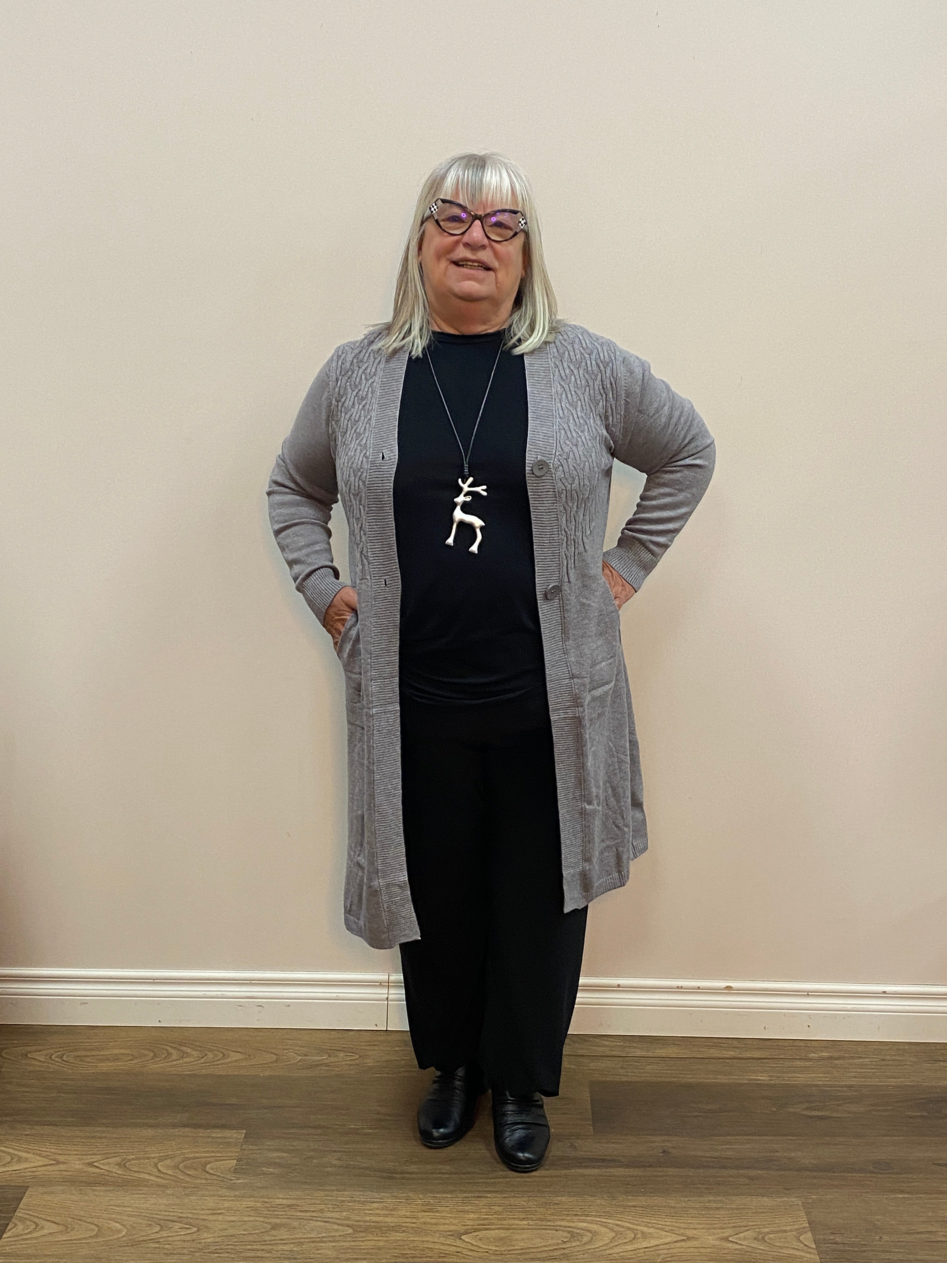 Person wearing a gray cardigan, black top, and black pants in a room with beige walls and wooden flooring.